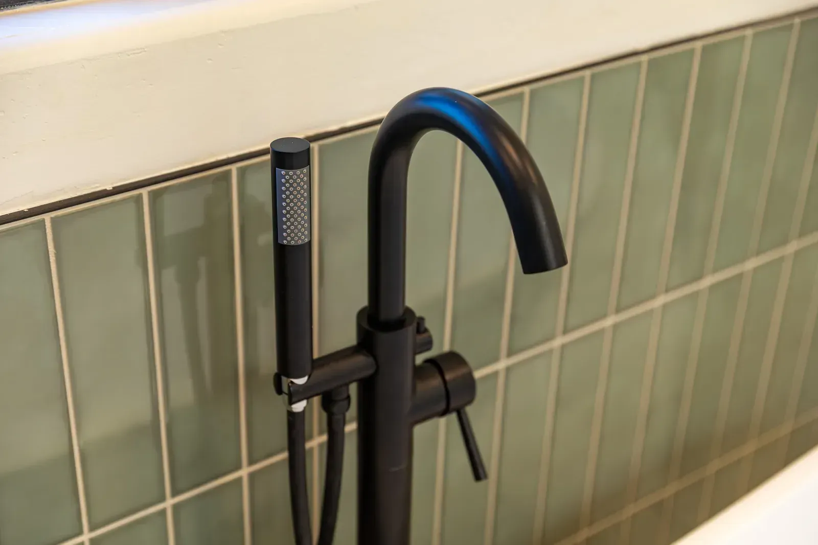 Black faucet with handheld sprayer mounted against green tile bathroom wall.
