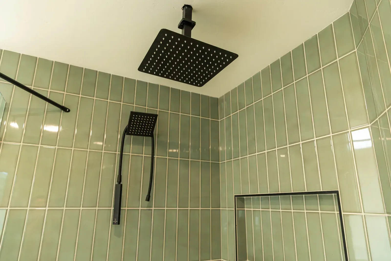 Green-tiled shower with two black showerheads and a metal grab bar