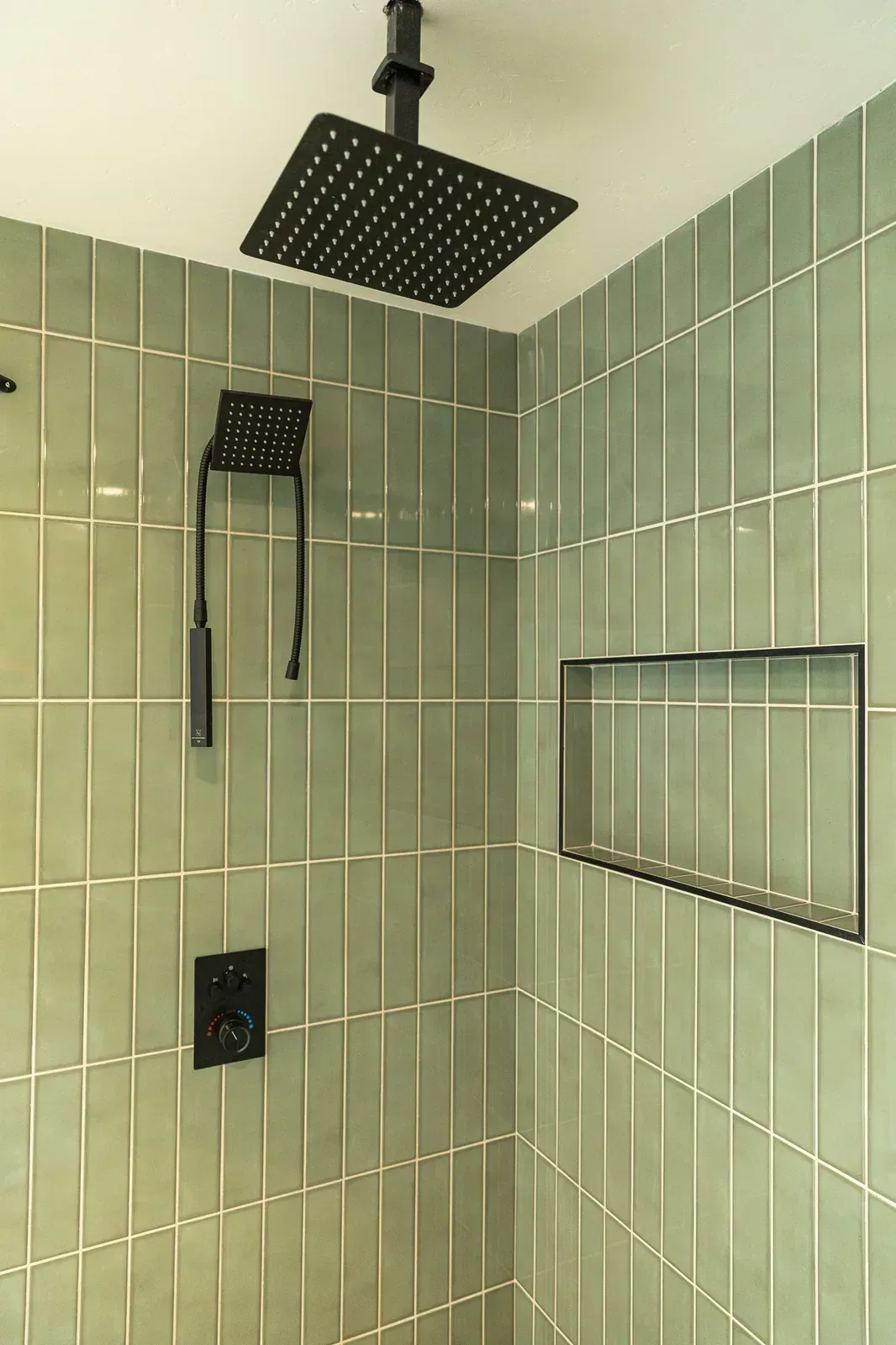 Green-tiled shower with black rain showerhead and handheld sprayer, plus recessed shelf and controls