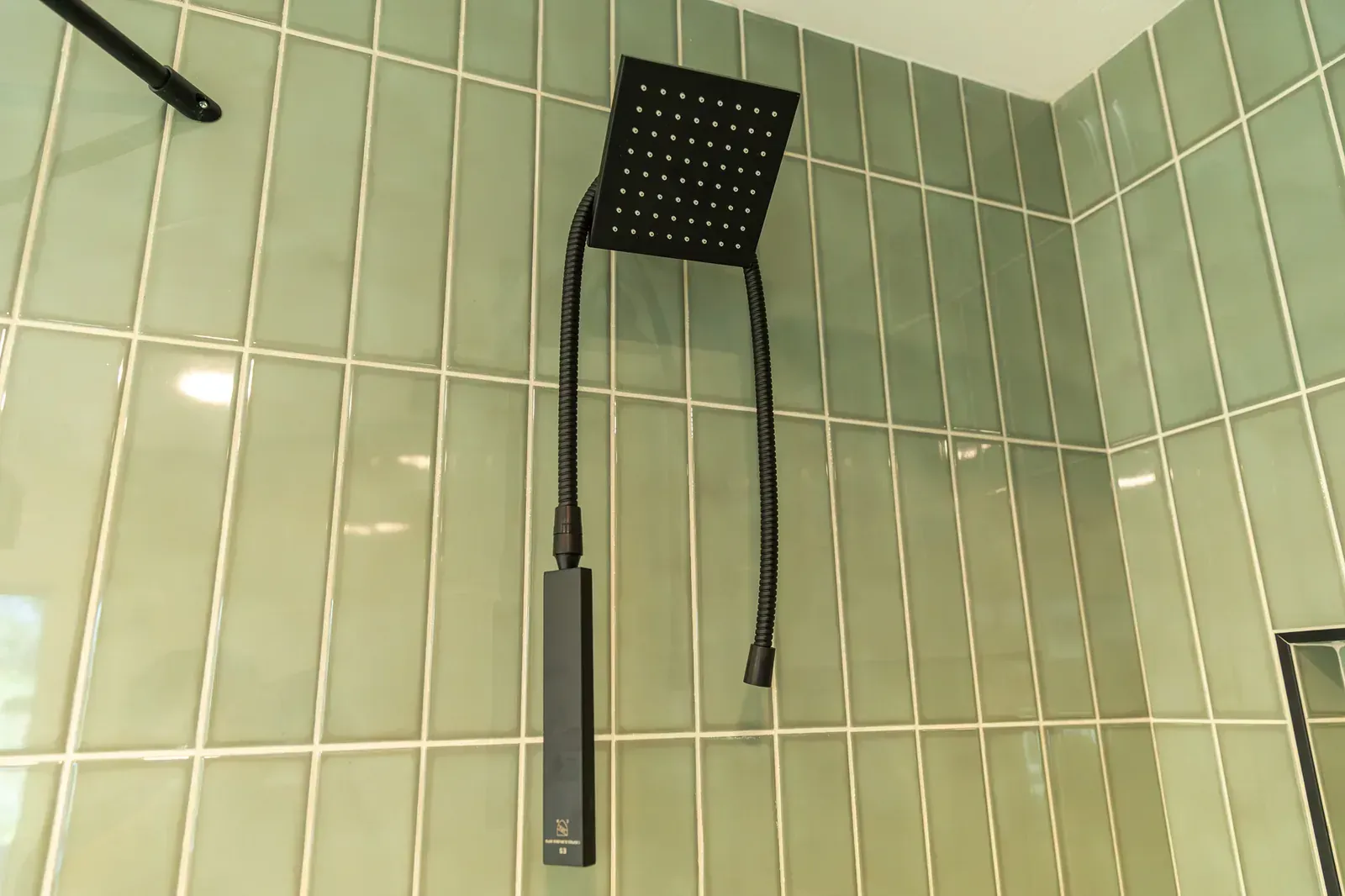 Black square shower head with handheld hose mounted on pale green tiled bathroom wall.