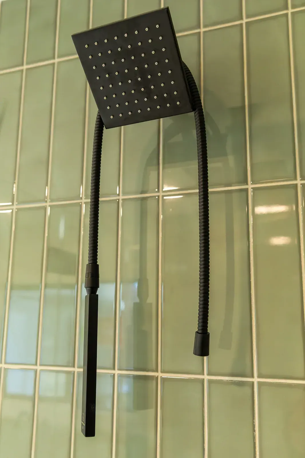 Black handheld shower head with hose hanging against green tiled bathroom wall