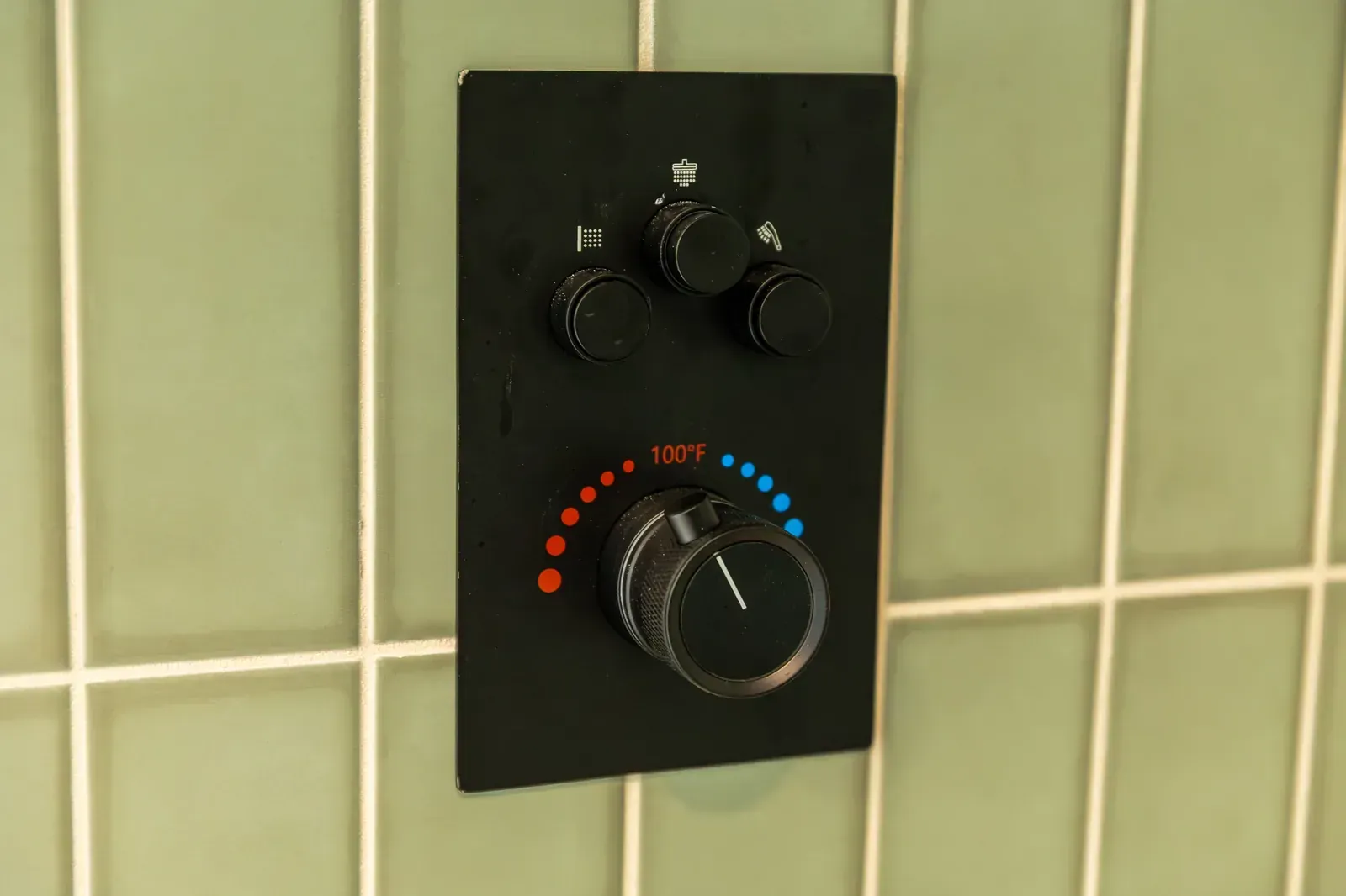 Black wall-mounted temperature control knob on green tiled wall with red and blue markings.