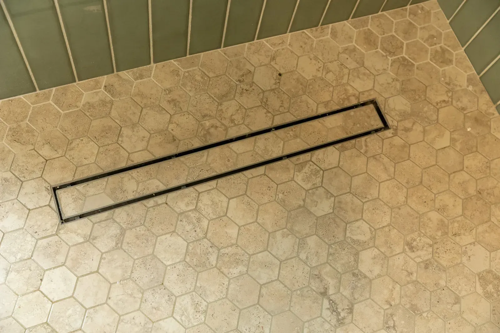 Bathroom floor with a long rectangular floor drain grate on beige hexagon tiles.
