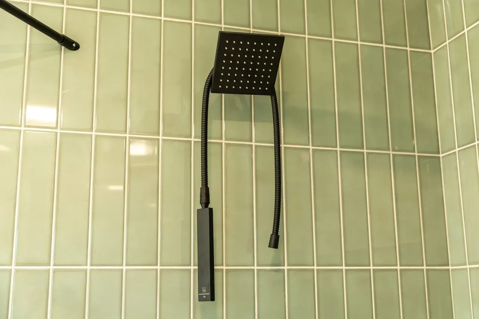Black showerhead and hose mounted on pale green tiled bathroom wall.