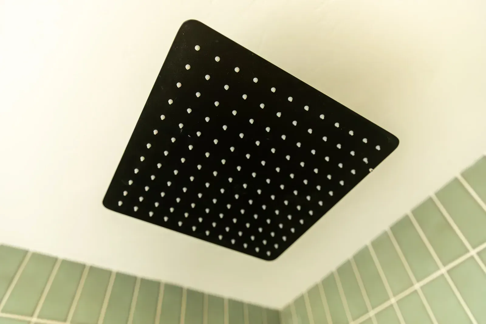 Black square rainfall showerhead on a white ceiling above green bathroom tiles