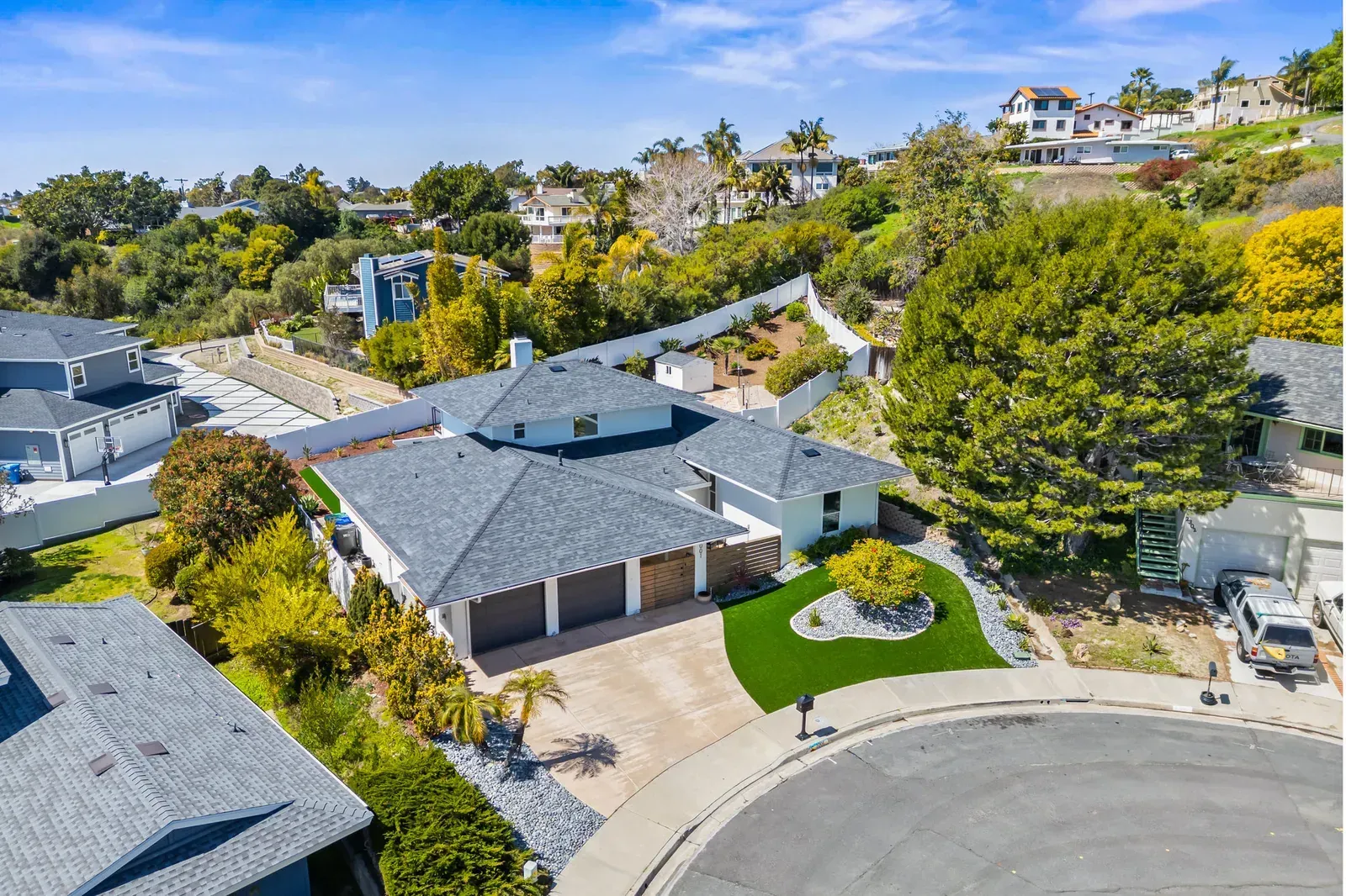 Aerial view of a suburban house with a curved driveway, green lawn, and hillside homes in the background