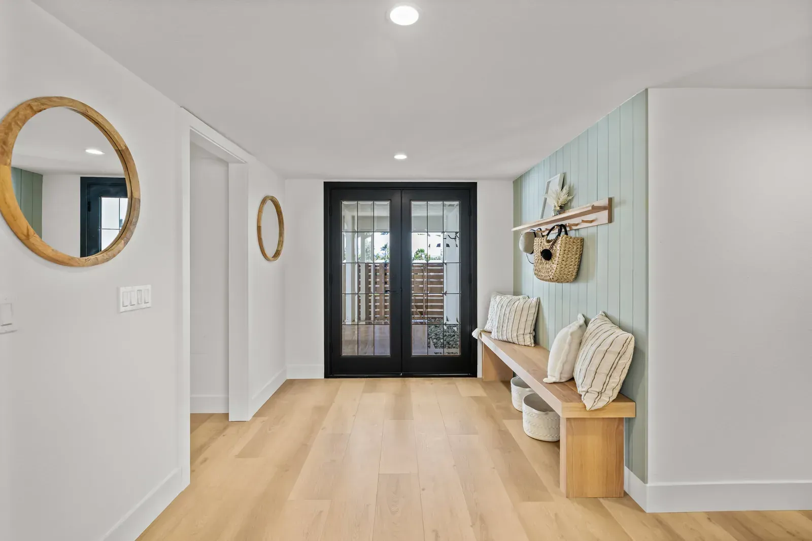 Bright hallway with light wood floors, round wall mirror, bench, and black glass doors at the end