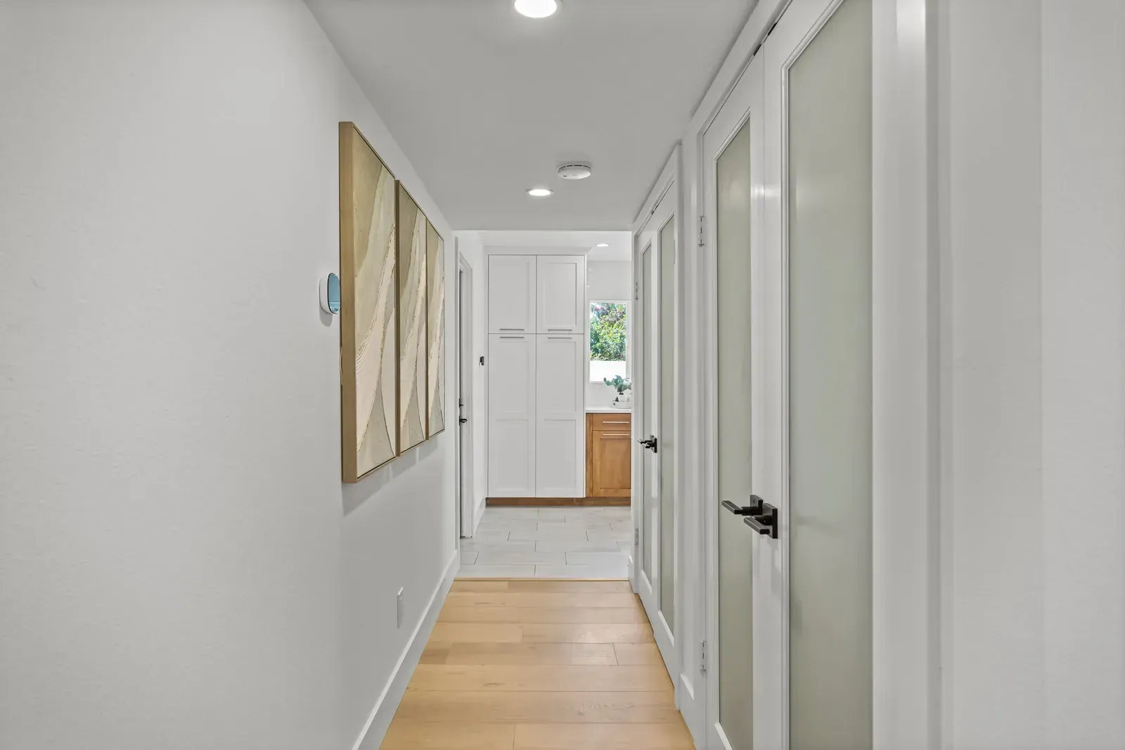 Bright hallway with white walls, wood floor, glass doors, and artwork leading to a kitchen.