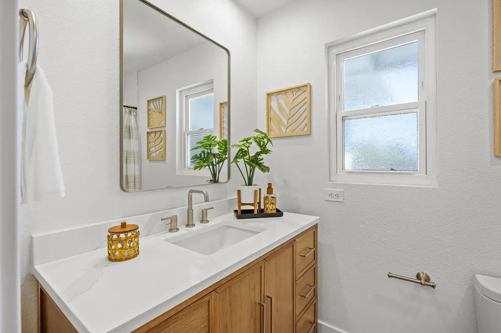 Bright bathroom vanity with wood cabinets, white countertop, mirror, window, and small plant decor.