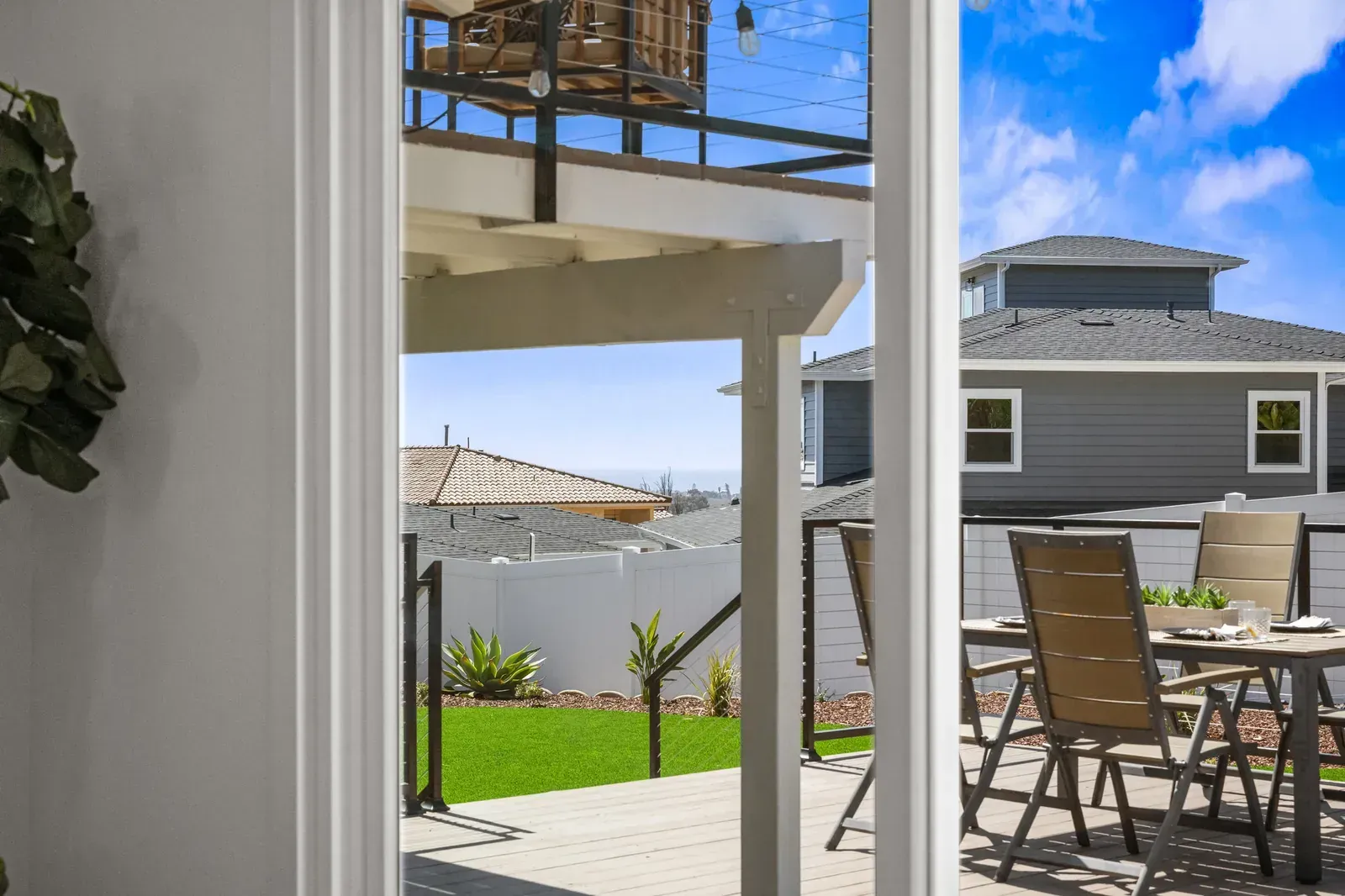 View through sliding doors to a sunny patio with table, chairs, green lawn, and neighboring house.