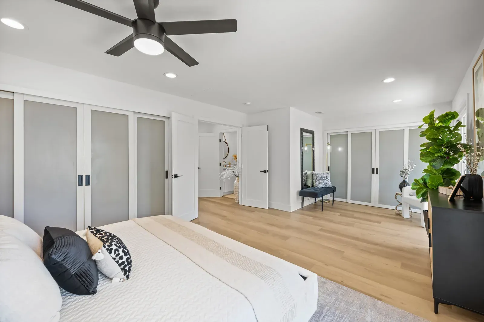 Bright bedroom with a bed, ceiling fan, frosted closet doors, wood floors, and a plant by the window.