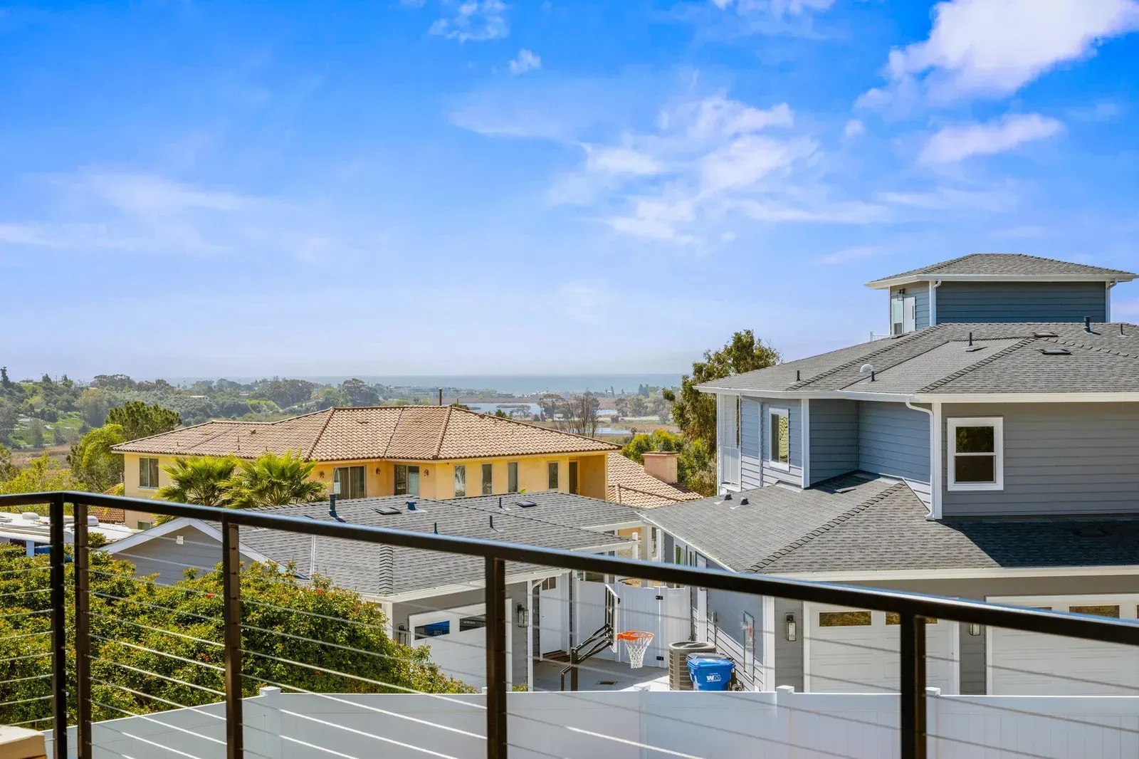 Balcony view of suburban rooftops, trees, and distant hills under a blue sky