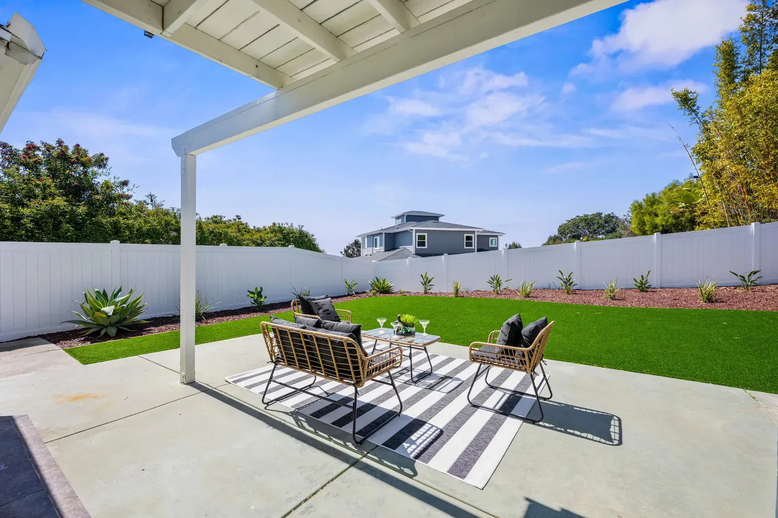 Covered patio with outdoor seating, white fence, green lawn, and blue sky in a backyard.