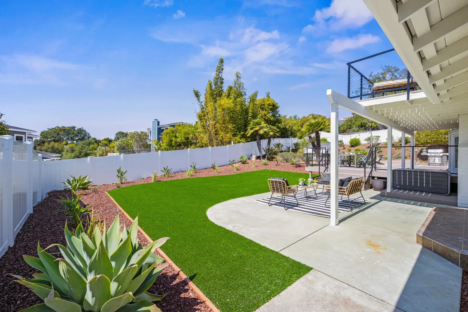 Backyard patio with artificial grass, stone path, and white fence under a covered deck
