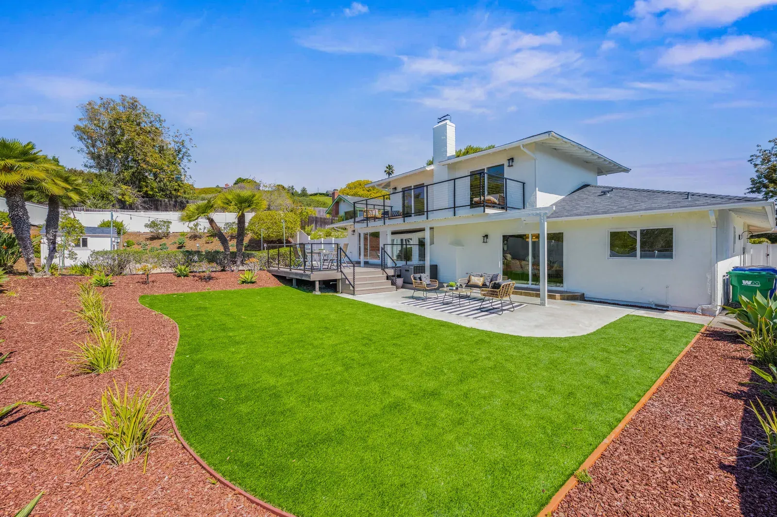 Modern white house with a bright green lawn, red mulch landscaping, and outdoor patio seating.