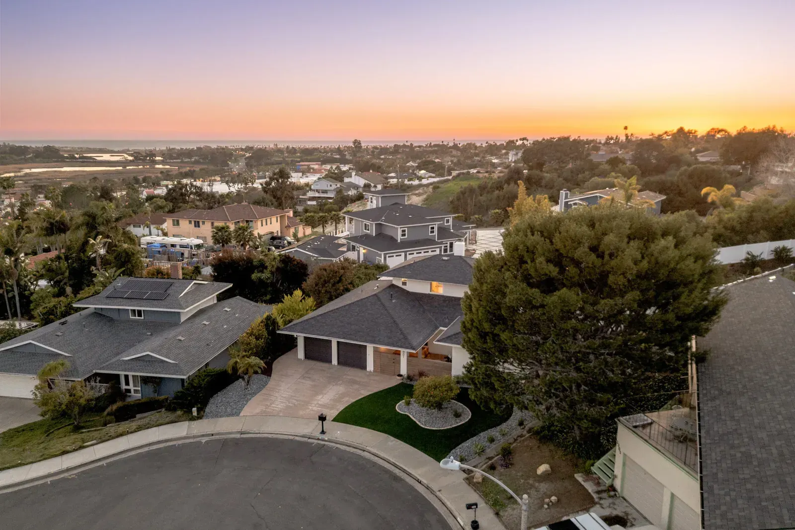 Suburban neighborhood at sunset with rooftops, winding streets, and distant city lights