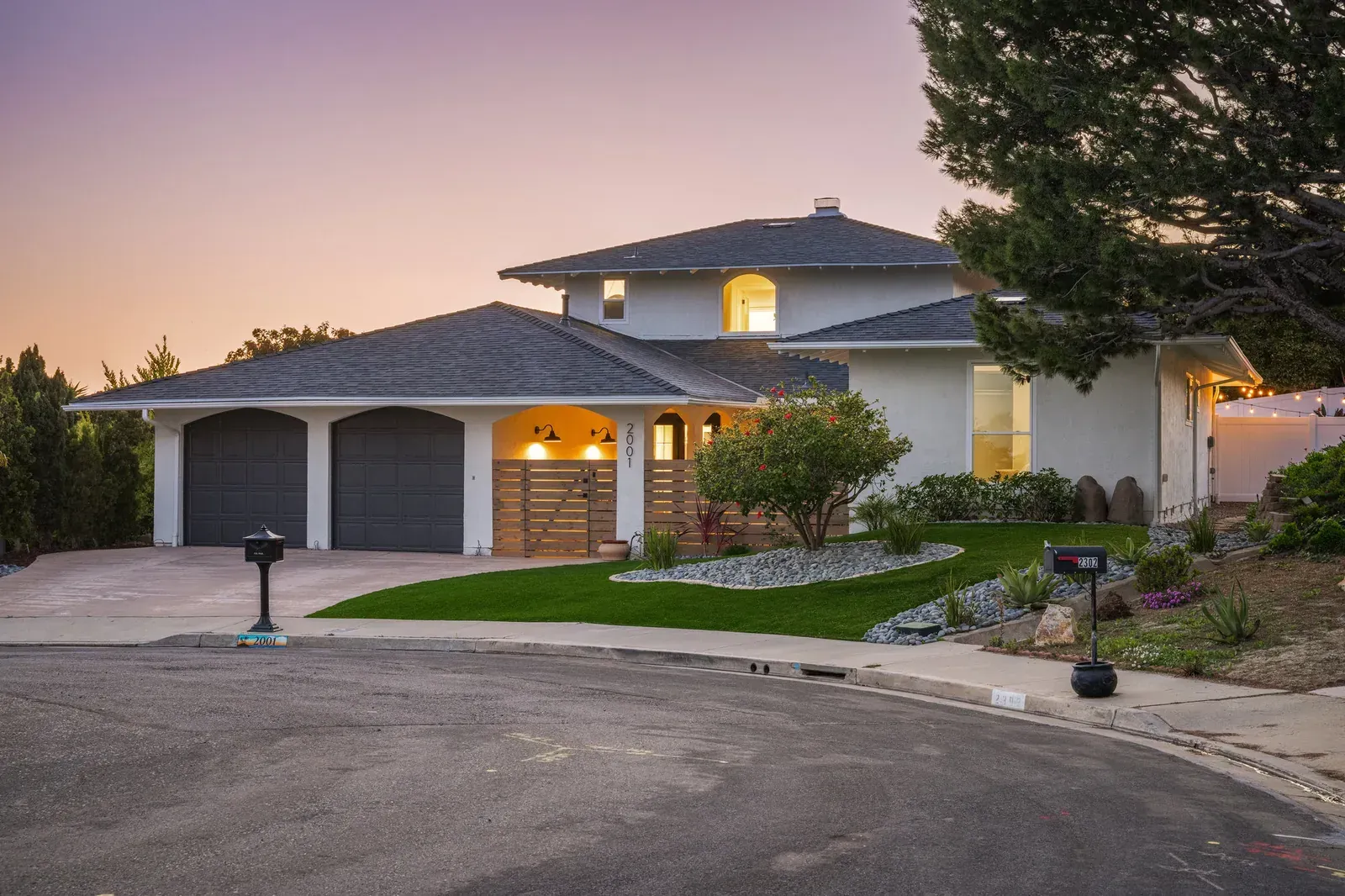 Suburban two-story house with lit windows at dusk, driveway, garage, and landscaped front yard