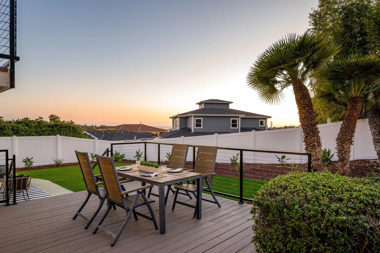 Patio with outdoor dining set overlooking a waterfront at sunset, framed by shrubs and palm trees