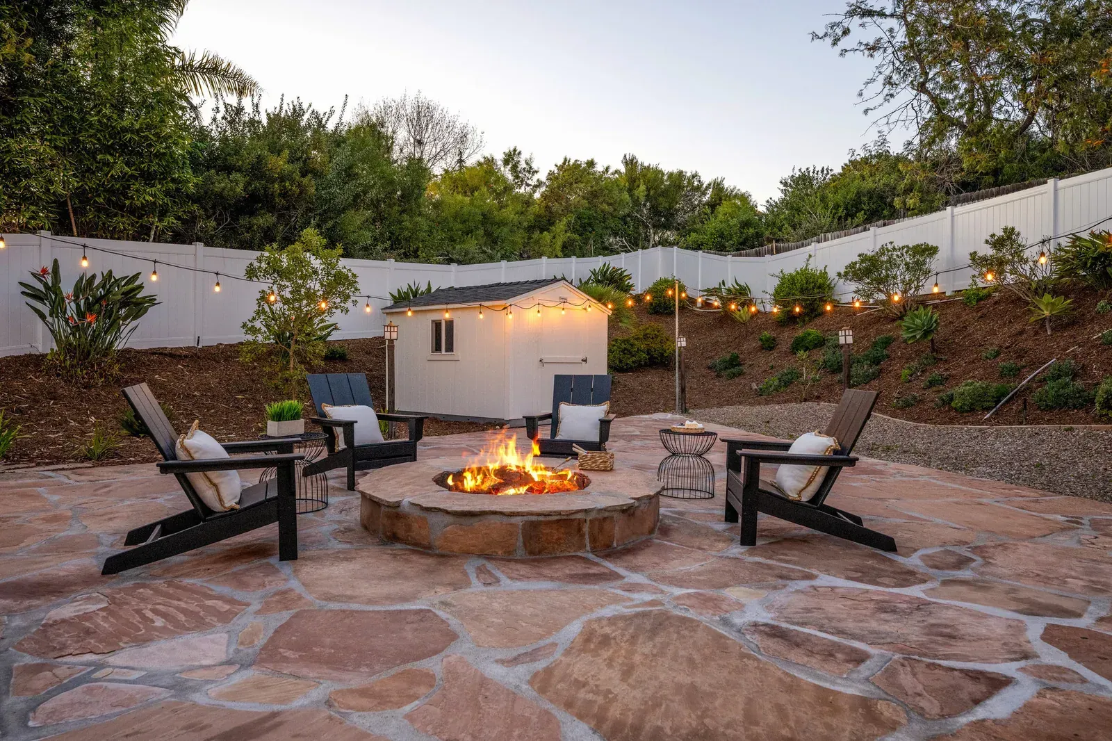 Backyard stone patio with fire pit and Adirondack chairs beside a small white outdoor fireplace