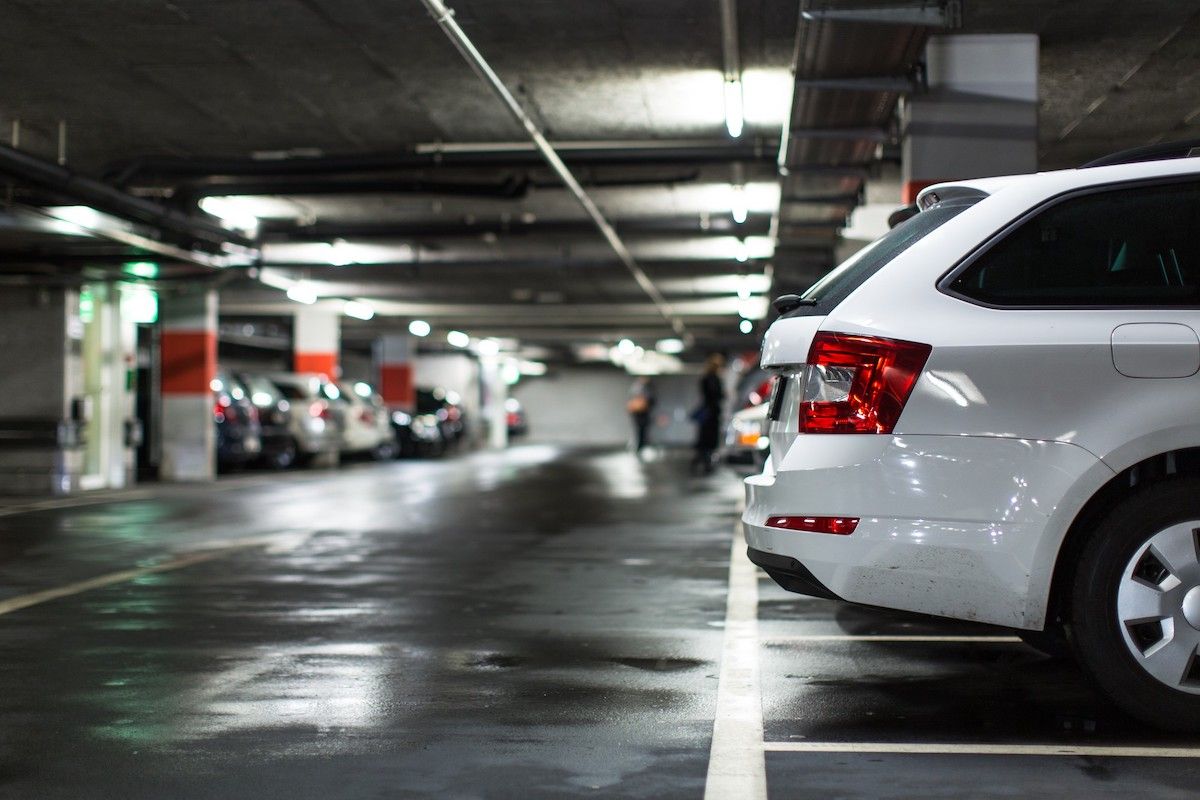 White car parked in a wet, dimly lit parking garage. Other cars and people are visible in the background.