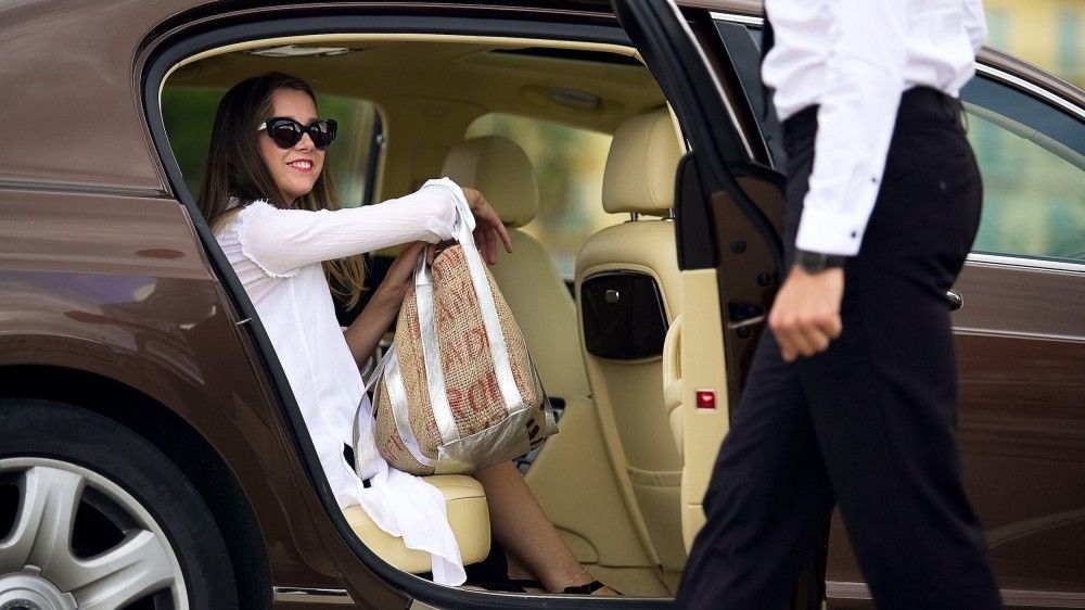 Woman in sunglasses smiles in a luxury car while a person in a suit opens the door.