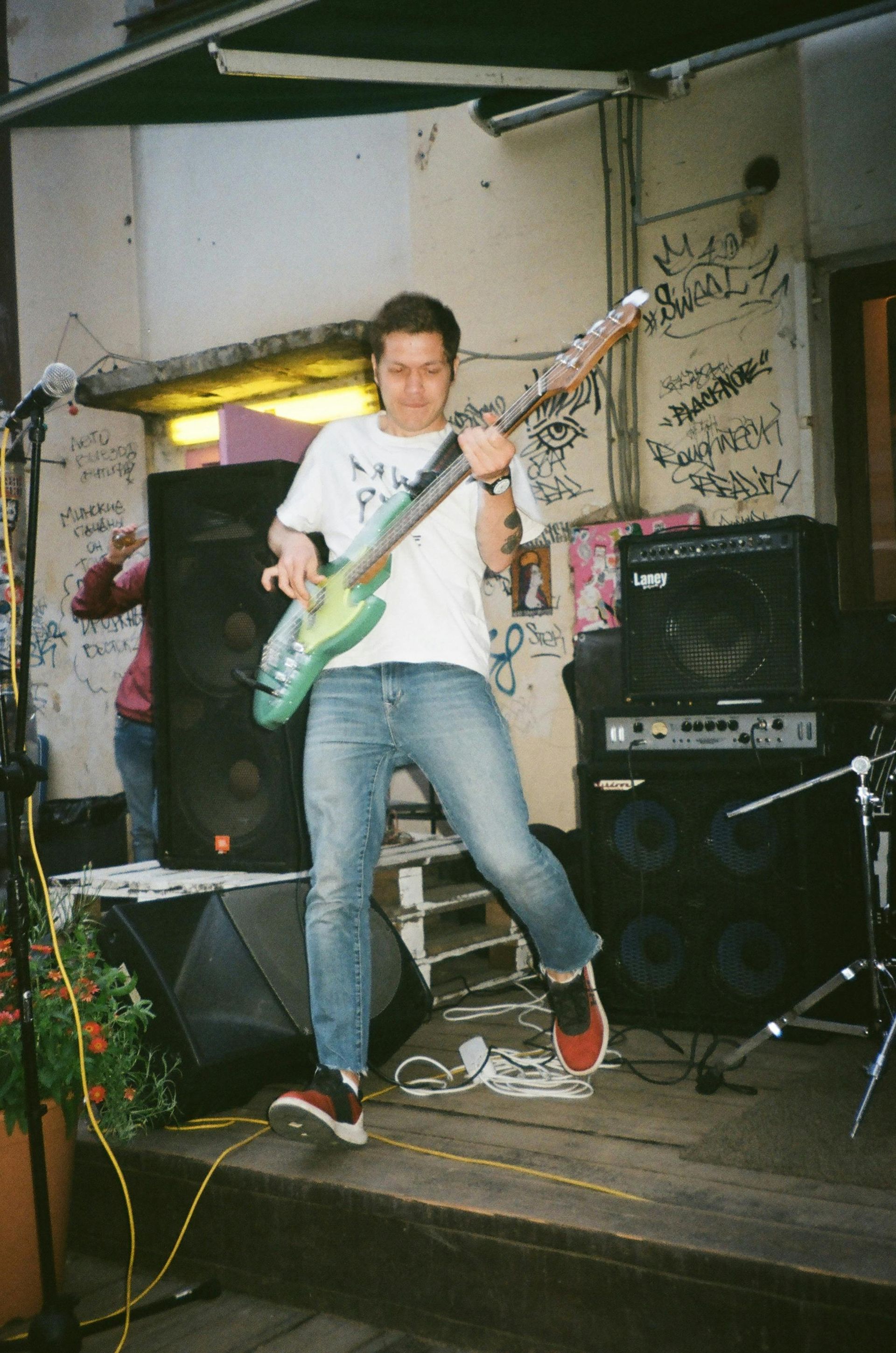 Man speelt basgitaar op een podium, draagt een wit T-shirt, spijkerbroek en rode schoenen. Buiten.