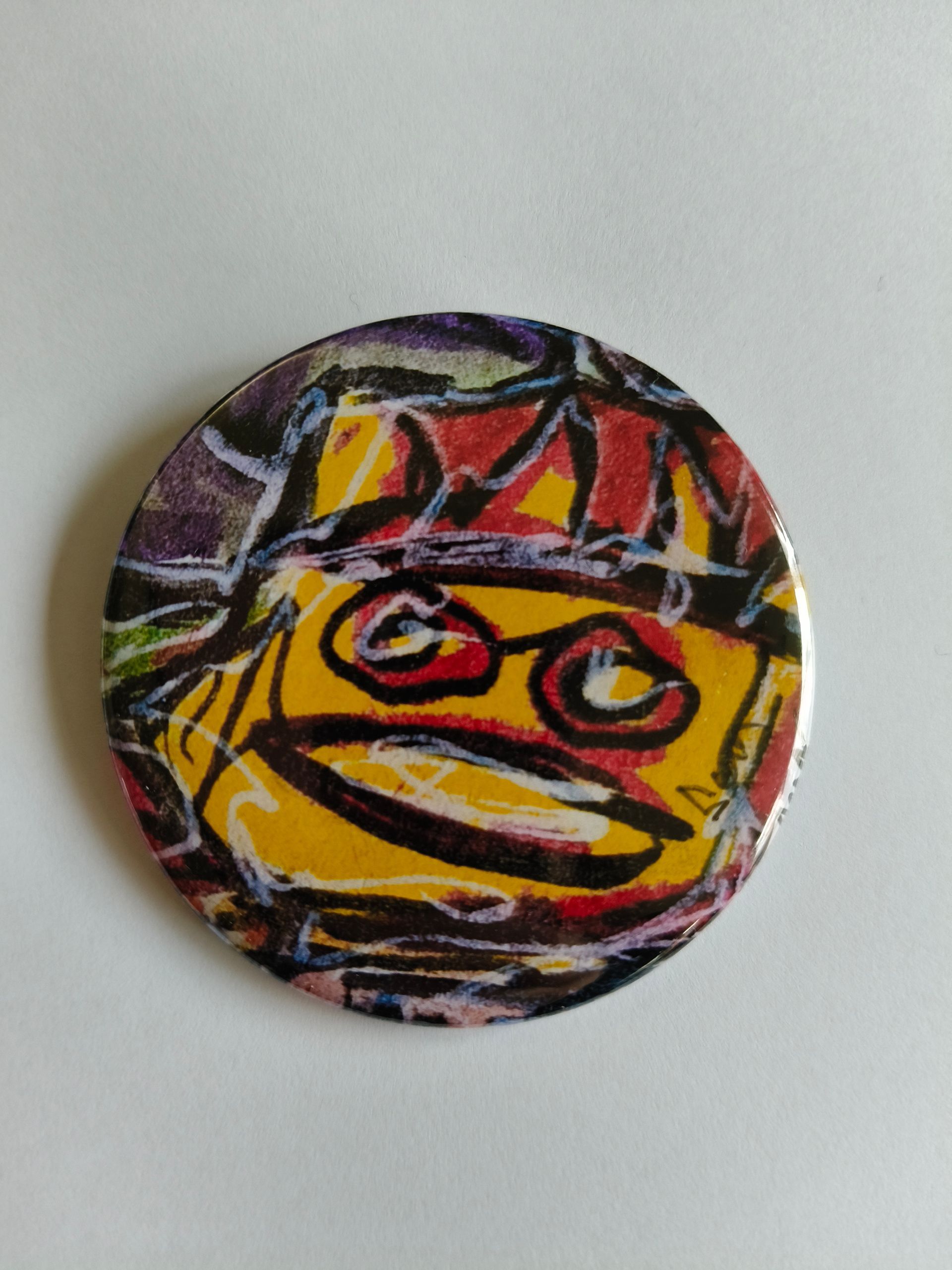 Een ronde button met een gestileerd, geel cartoongezicht met donkere, krabbelige contouren, grote ogen en een brede mond.