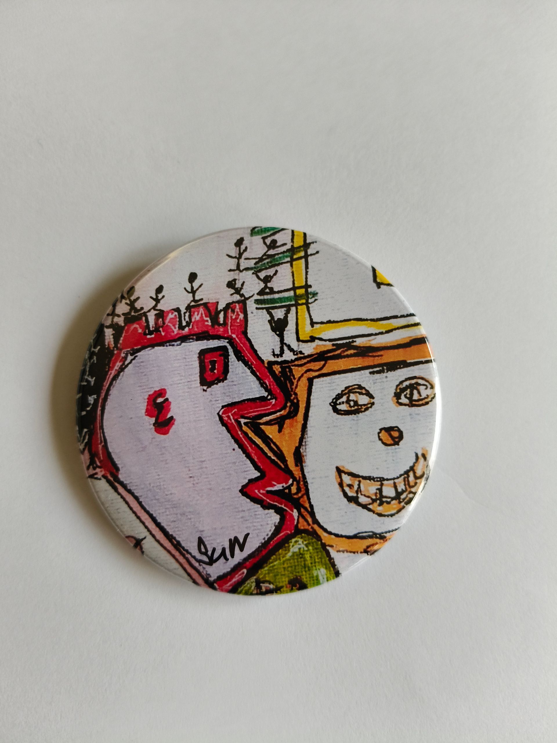 Een ronde knop met een kleurrijke, abstracte tekening van twee gezichten en kleine figuren tegen een witte achtergrond.