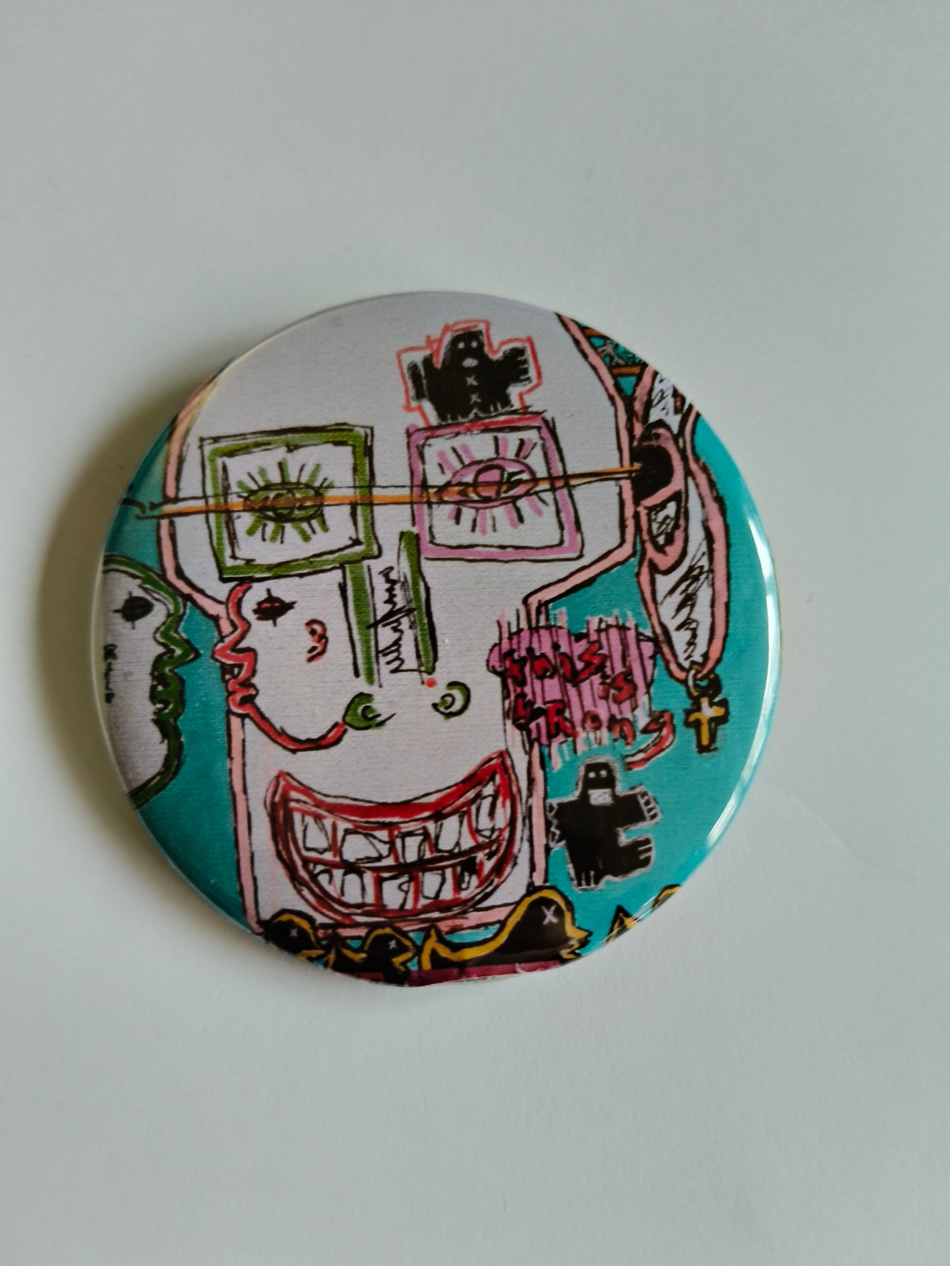Een ronde speld met een kleurrijk, graffiti-achtig portret met overdreven ogen, brede grijns en een turquoise achtergrond.
