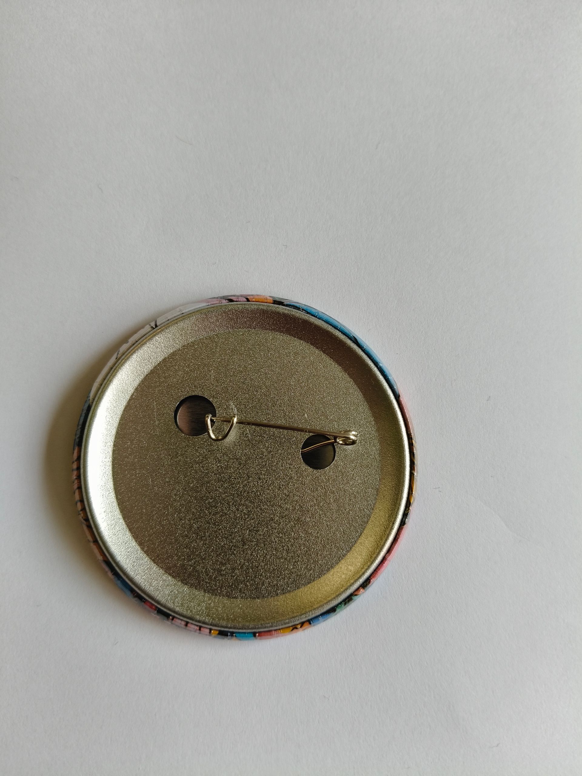 Een ronde button met een gestileerd, geel cartoongezicht met donkere, krabbelige contouren, grote ogen en een brede mond.
