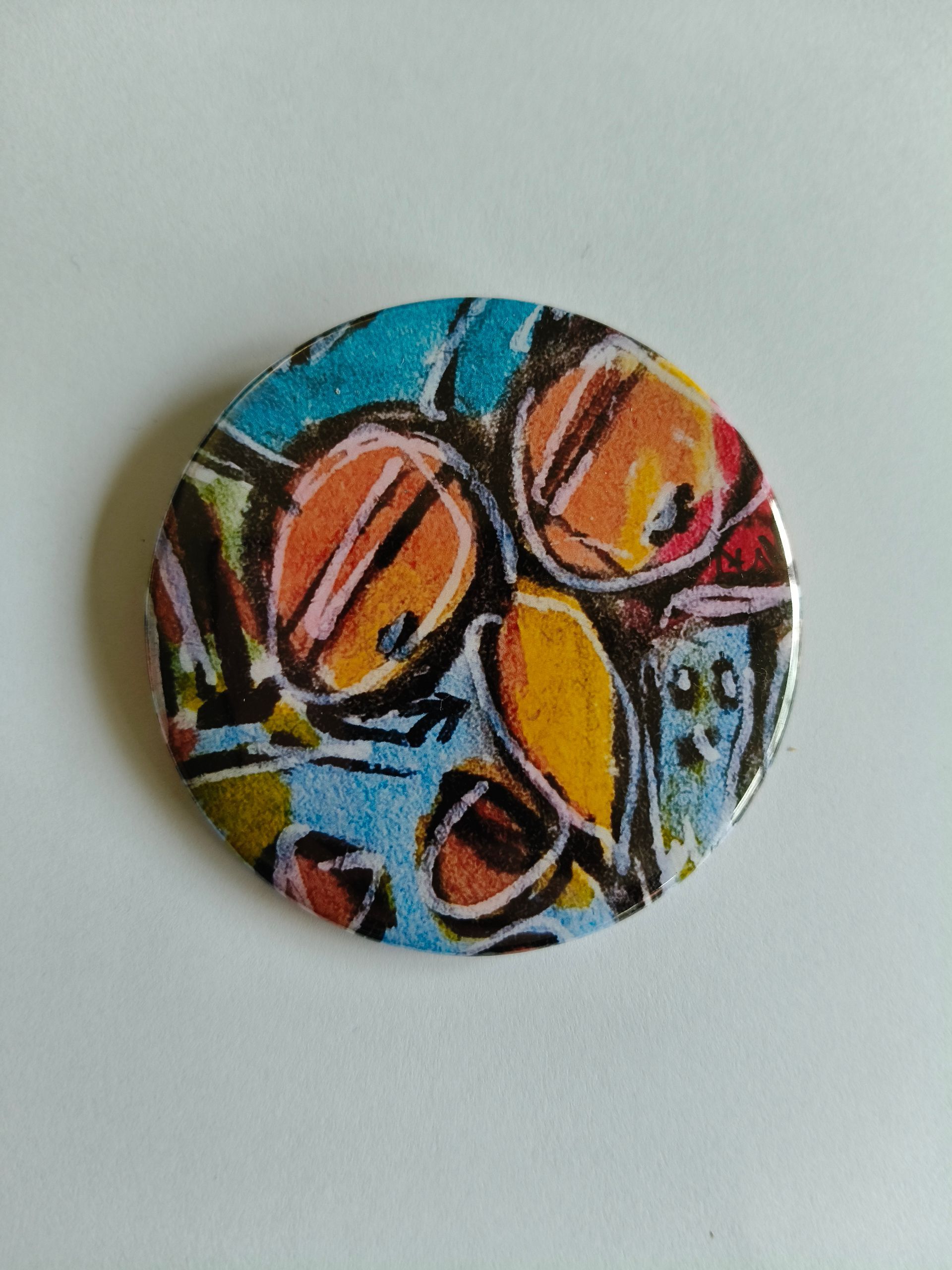 Een ronde speld met een abstract, kleurrijk patroon van overlappende oranje, gele en blauwe vormen met zwarte contouren.