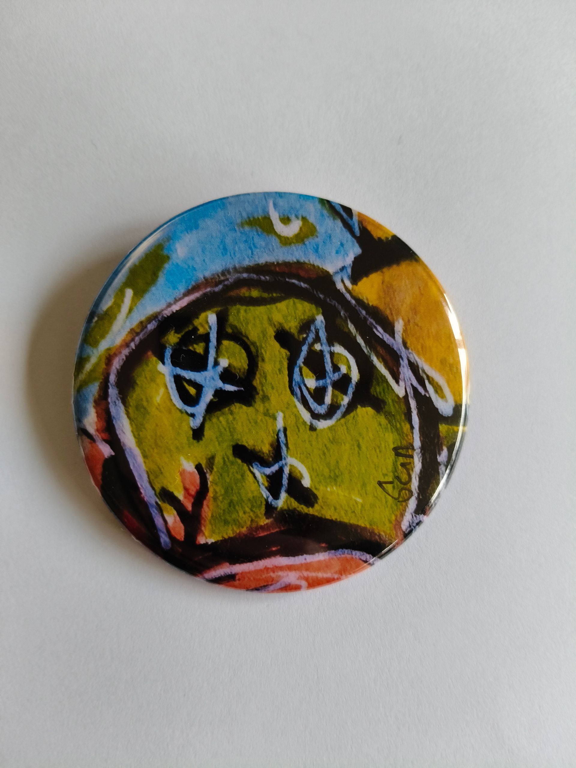 Een ronde button met een kleurrijke, abstracte tekening van een gezicht met doorgestreepte ogen en gekrabbelde details.