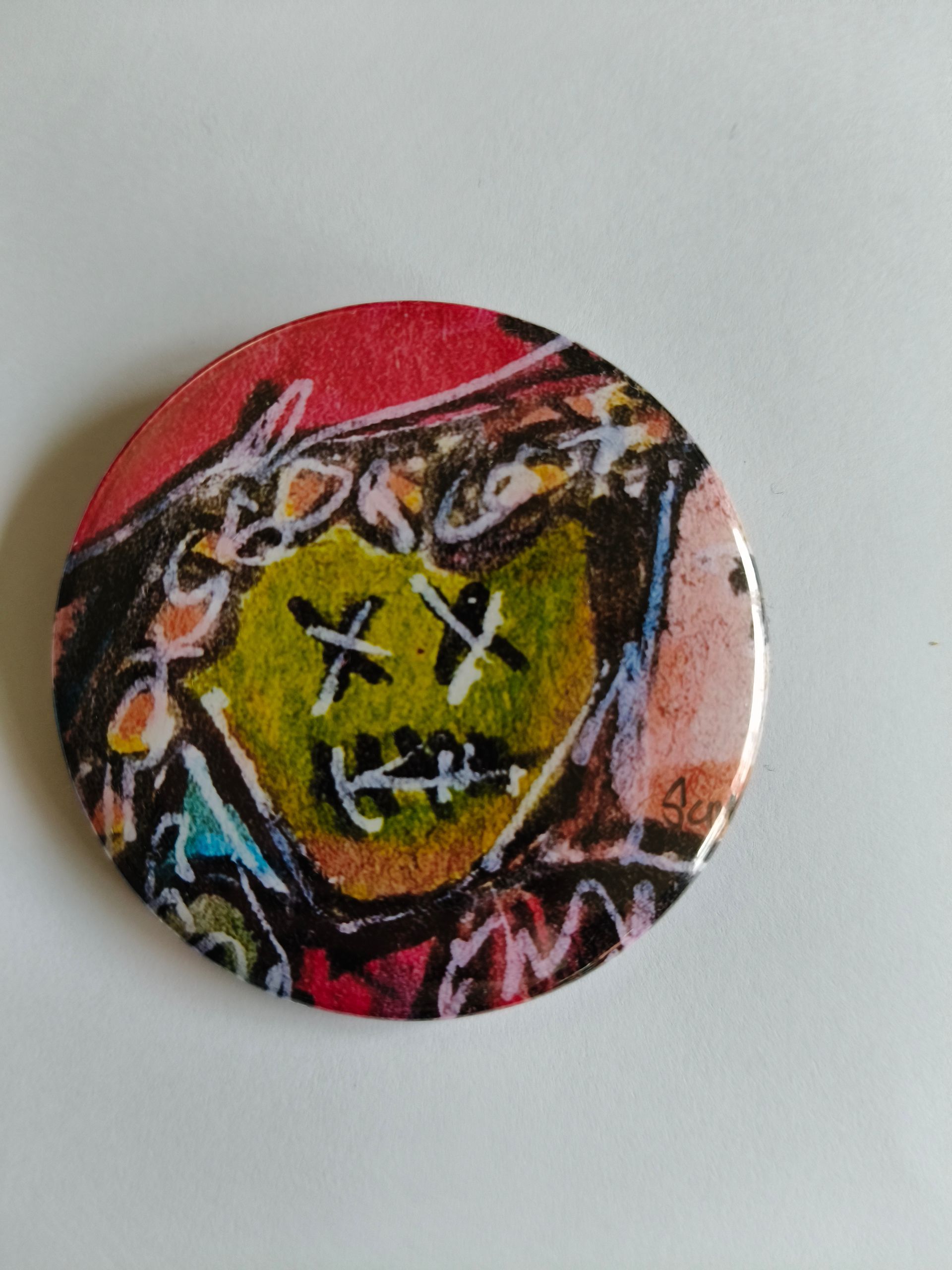 Een ronde knoop met een kleurrijke, abstracte tekening van een gezicht met 'X'-ogen en een gestikte mond.