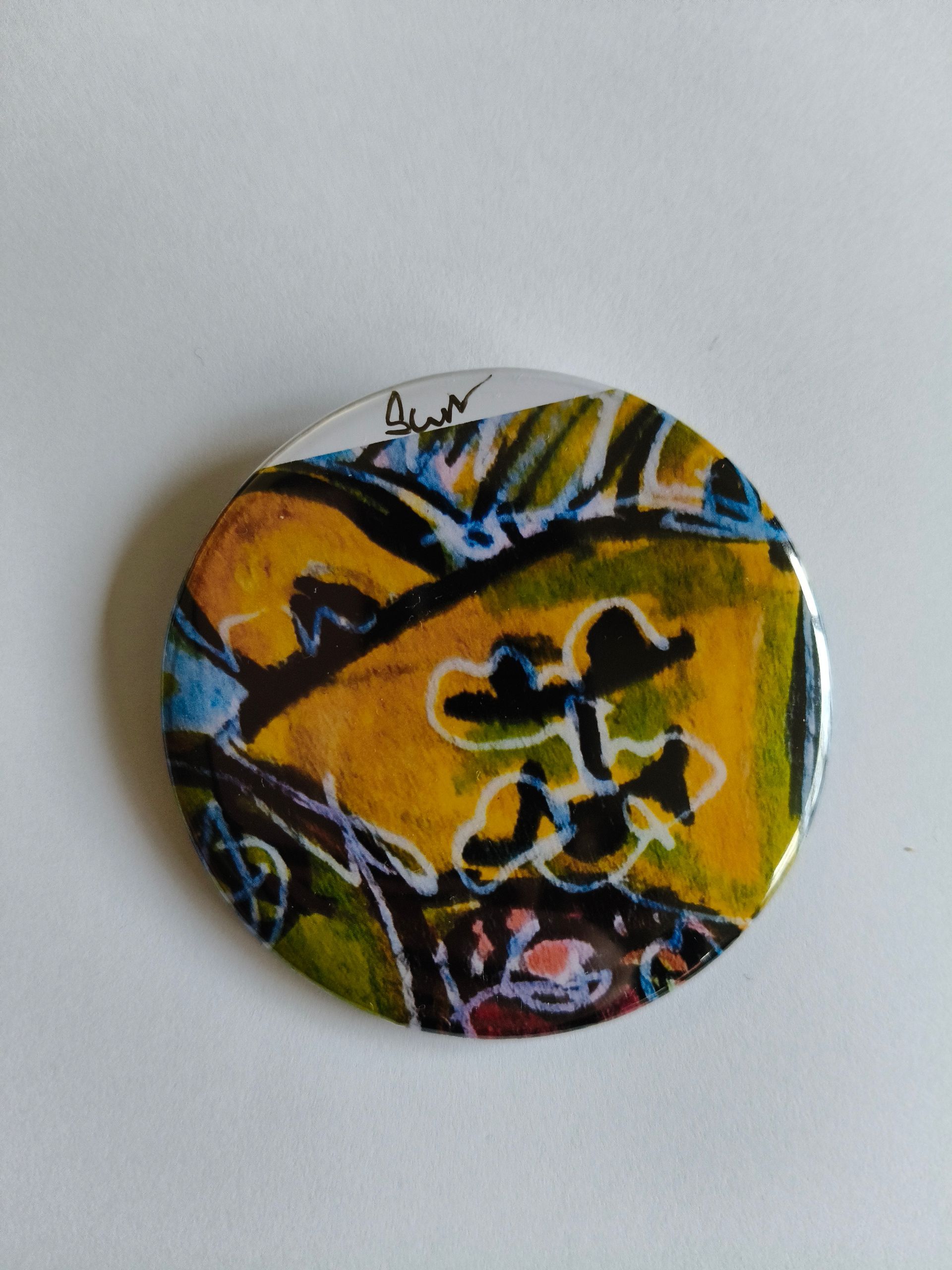 Een ronde speld met abstracte, expressieve penseelstreken in tinten geel, groen, blauw en zwart op een witte achtergrond.