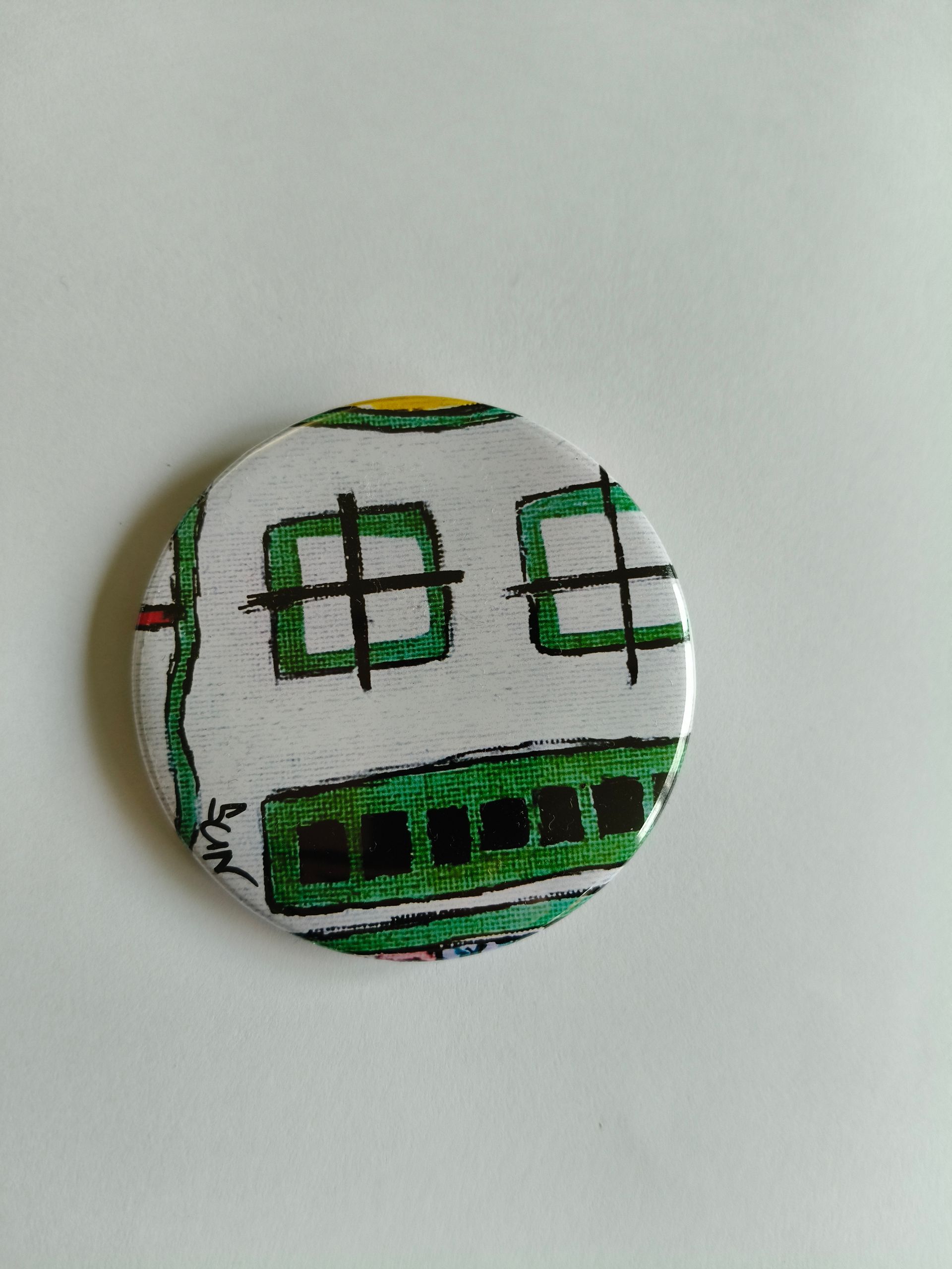 Een ronde knoop met een handgetekend, abstract groen gezicht met twee rechthoekige ogen en een brede, gesegmenteerde mond.