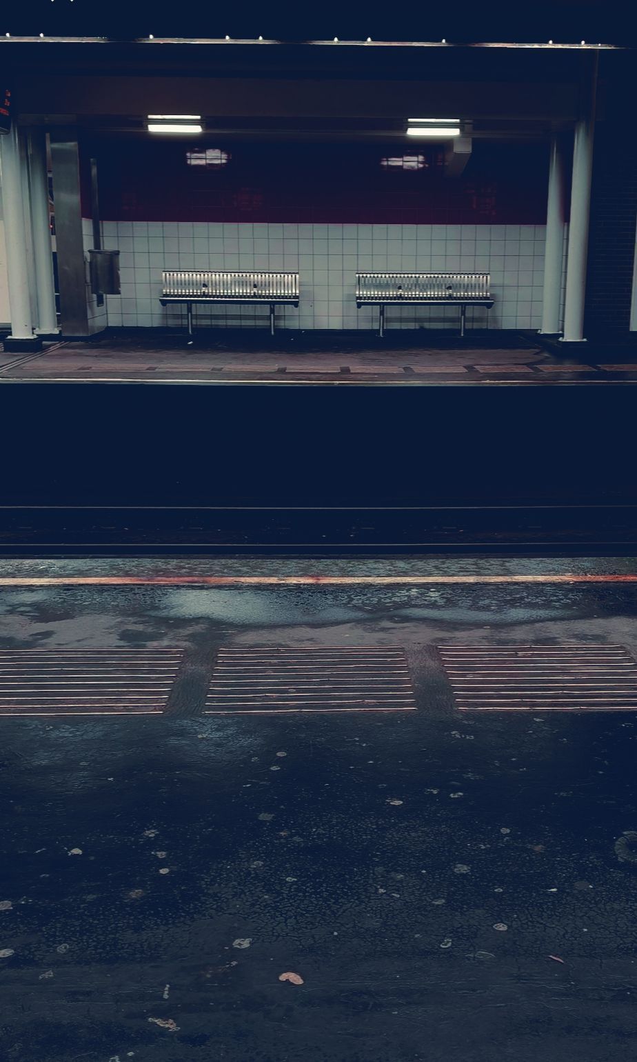 Een uitzicht over de spoorlijn naar een leeg metrostation met twee bankjes tegen een betegelde muur.