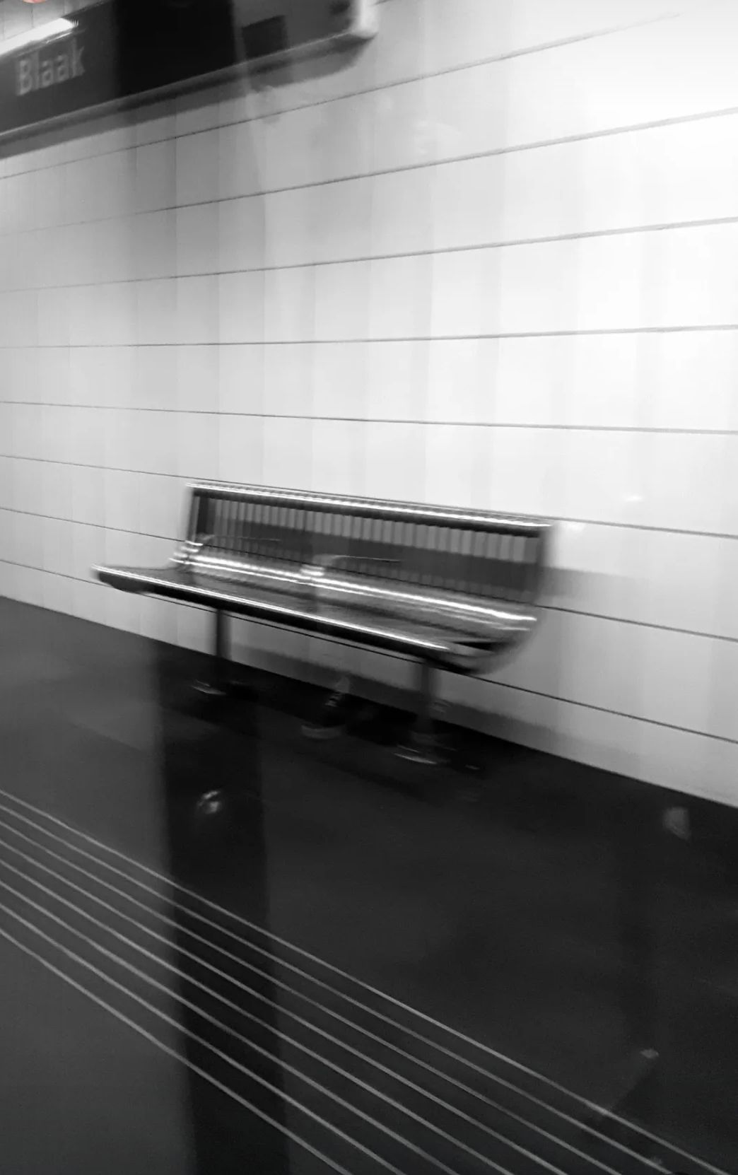 Een bewegingsonscherpe zwart-witfoto van een metrostationperron met een bankje tegen een betegelde muur.