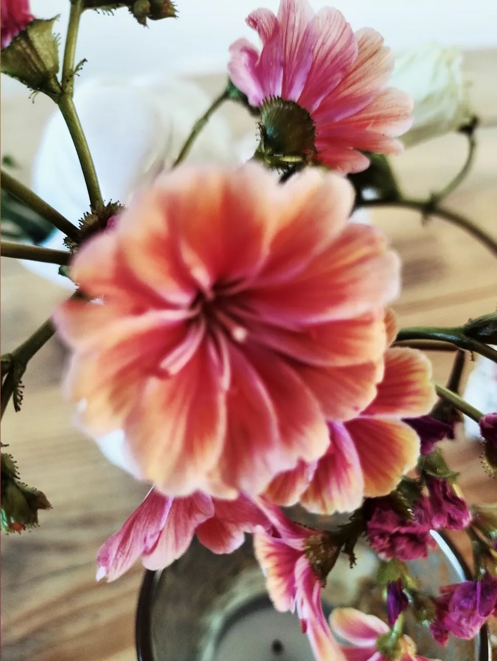 Een close-up van een boeket roze en perzikkleurige bloemen in een glazen vaas op een houten ondergrond.