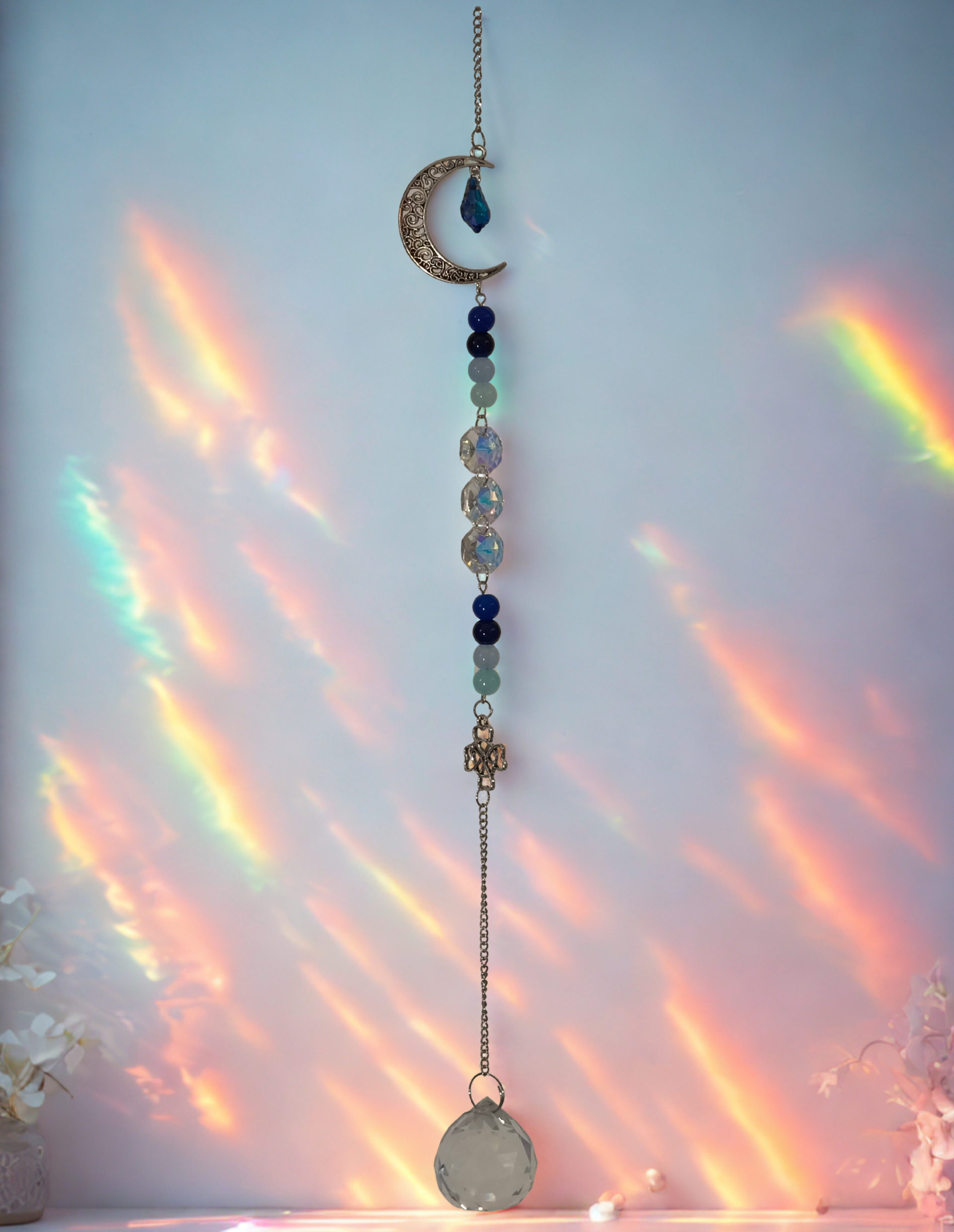 Een hangende raamdecoratie met een maan, blauwe en groene kralen en een kristallen traan, geplaatst tegen een muur