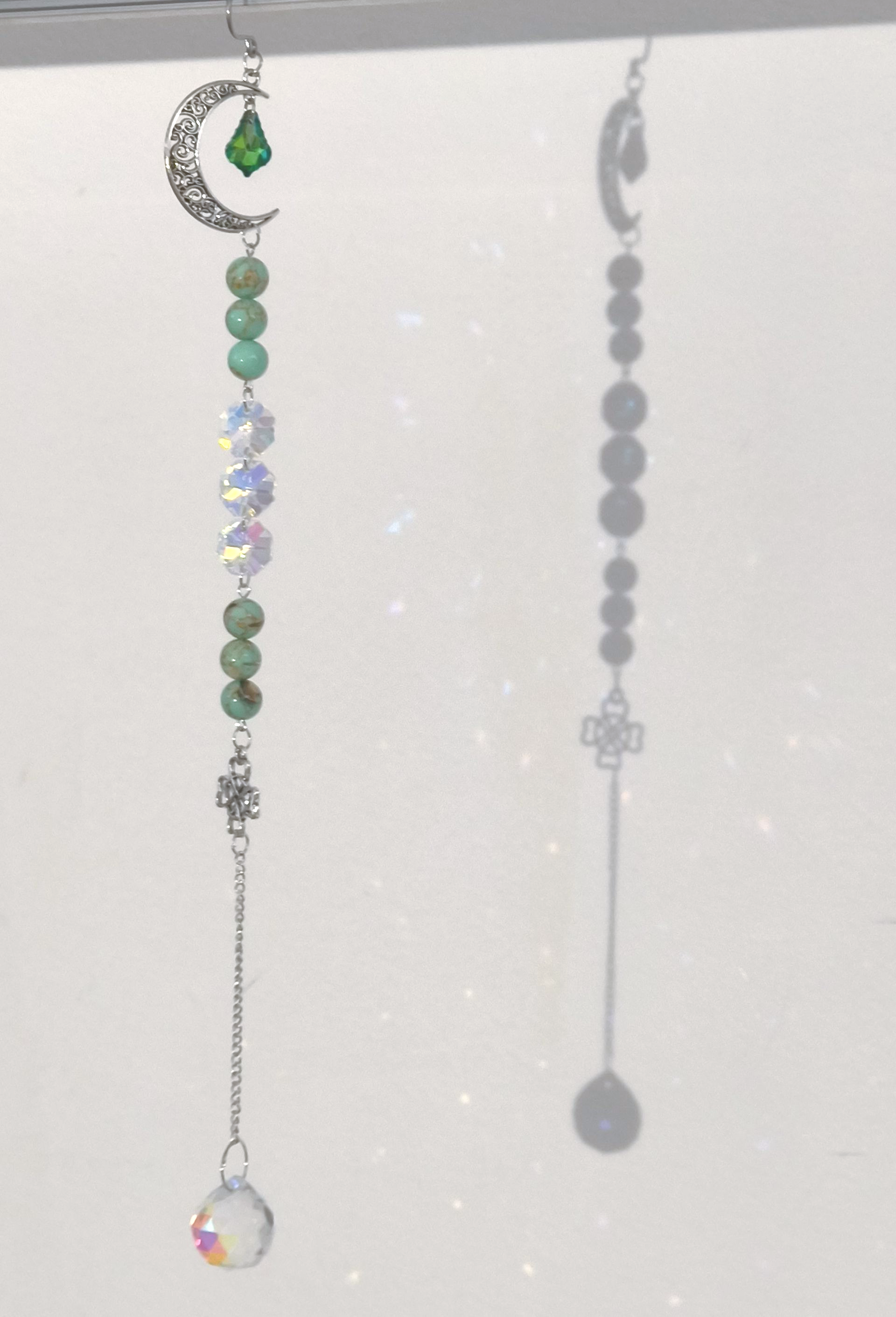 Een raamdecoratie-ornament met een zilveren halve maan, groene kralen, heldere iriserende kristallen en een glazen prisma.