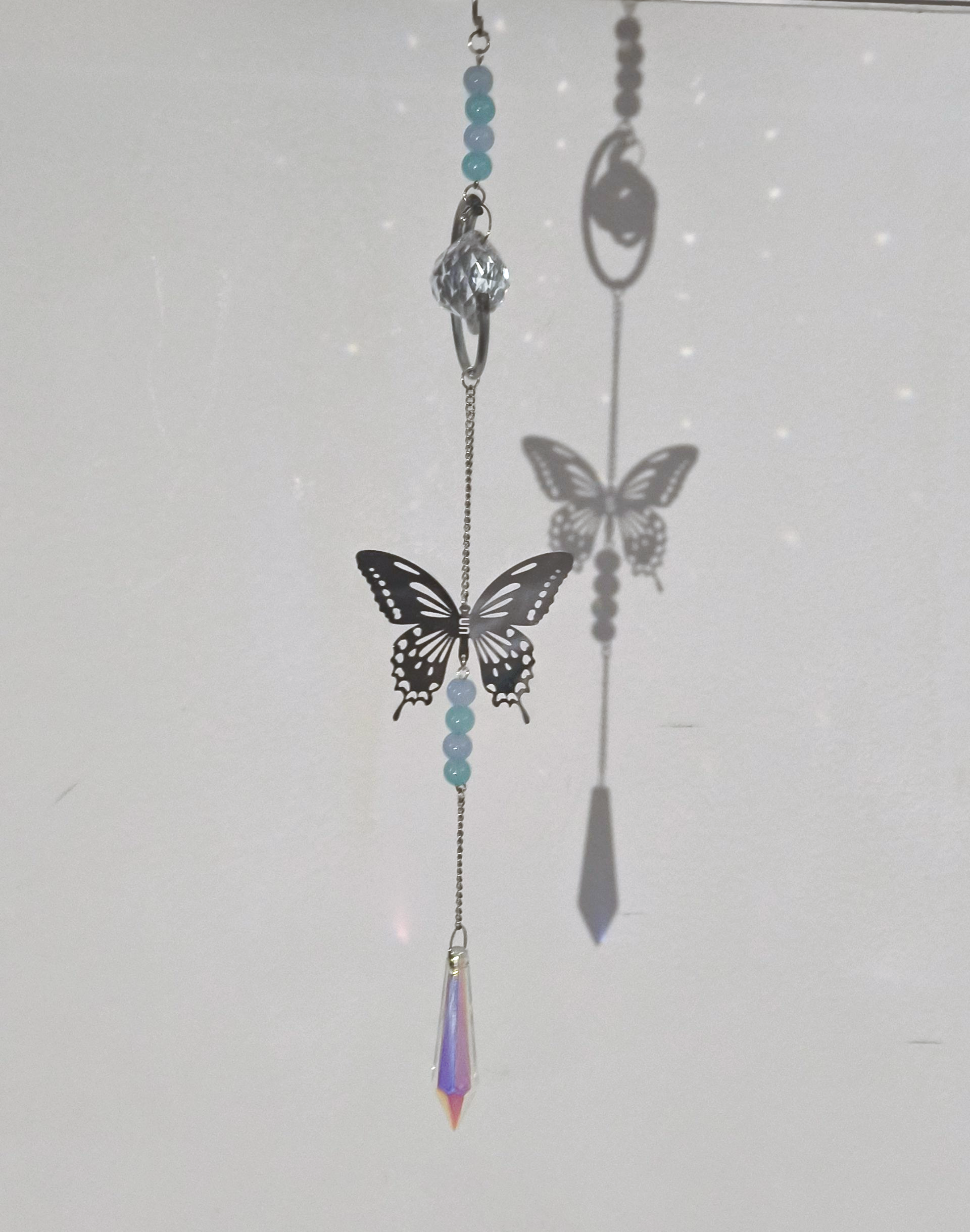 Een ornament met een metalen vlinder, lichtblauwe kralen, transparante kristallen bol en een lavendelkleurige prismahanger.