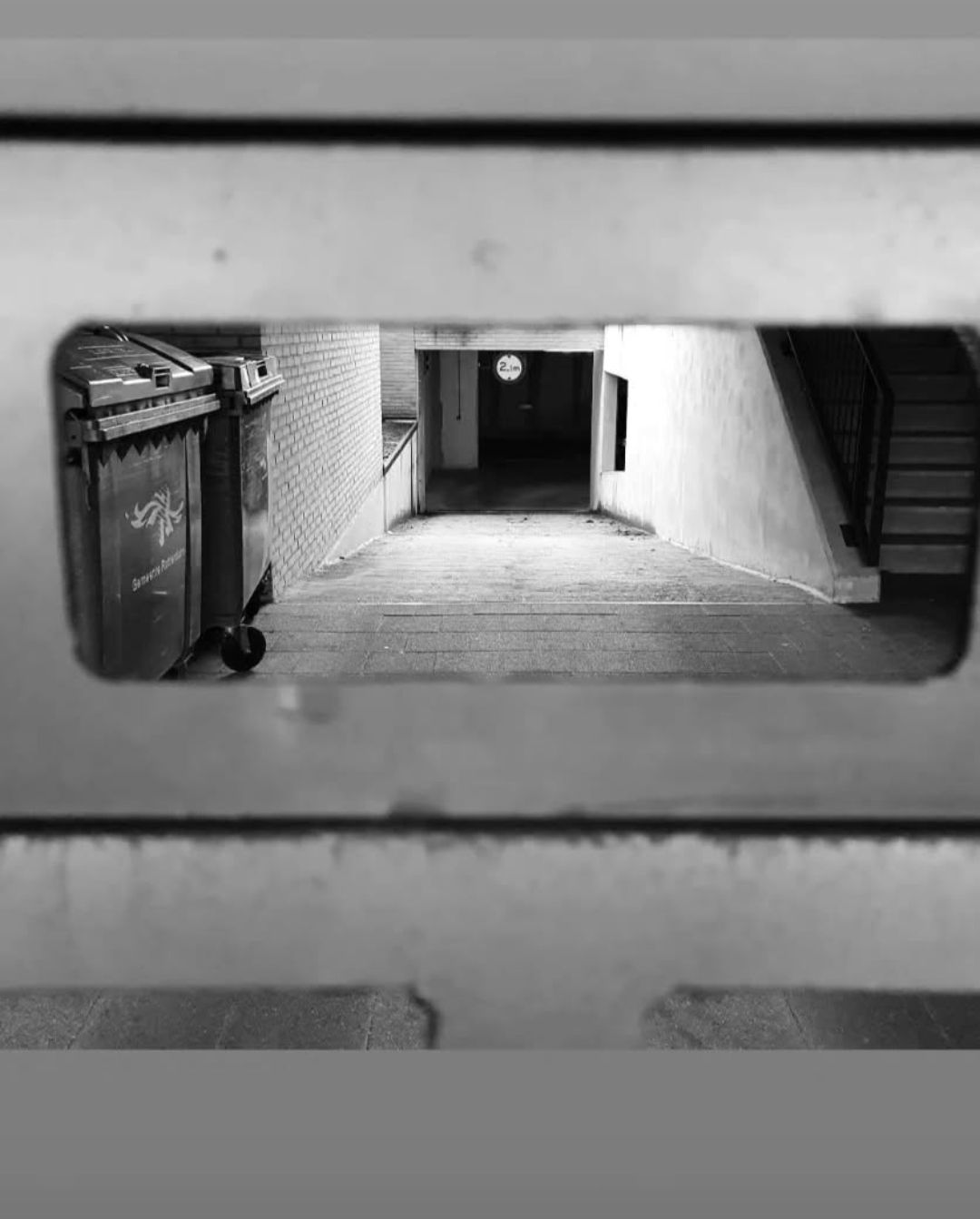 Een zwart-witbeeld door een rechthoekige uitsparing, waarop een donkere steeg met vuilnisbakken en een trap te zien is.