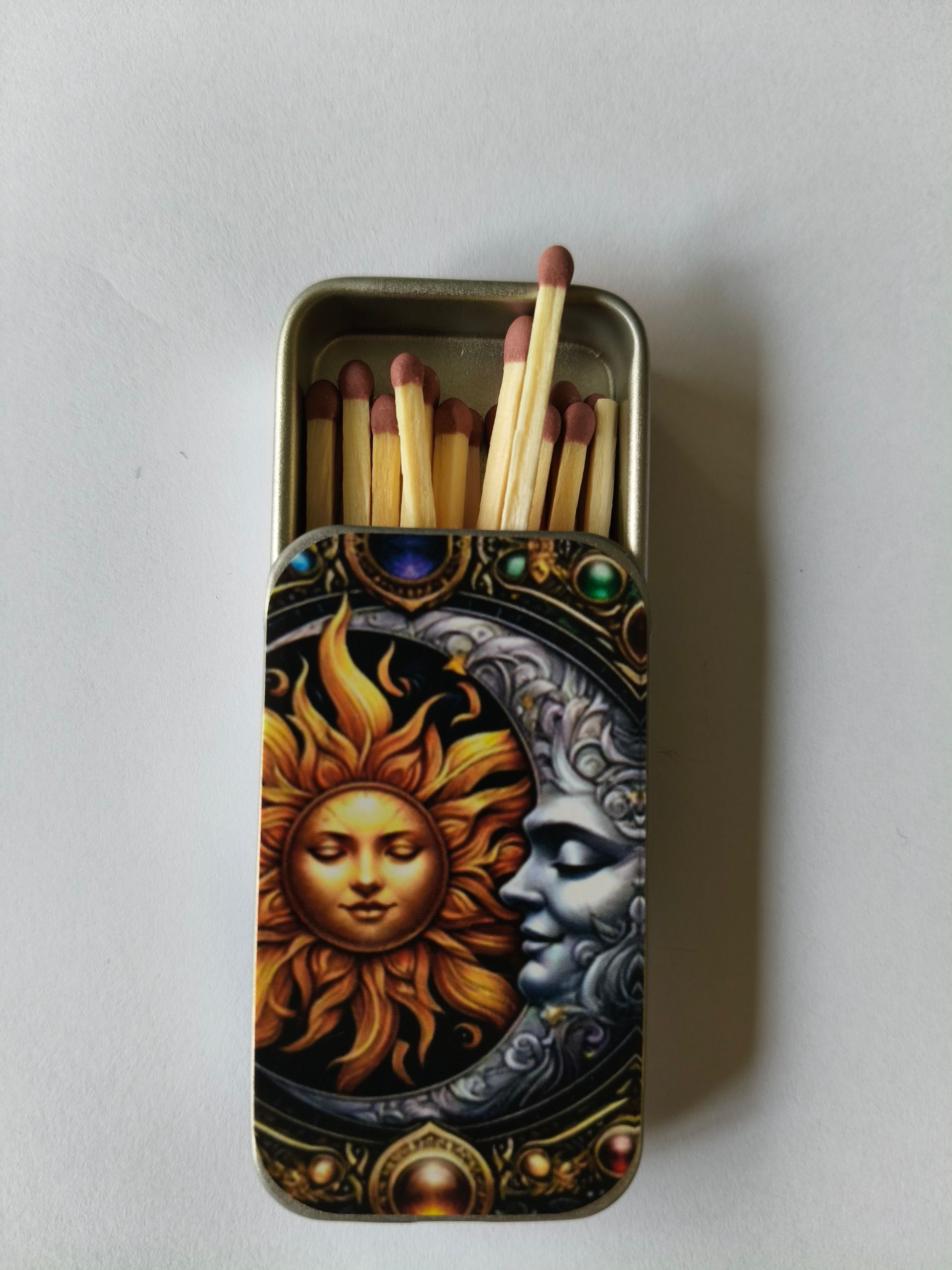Een metalen blikje met lucifers, met een decoratief zon- en maanmotief op het deksel tegen een witte achtergrond.