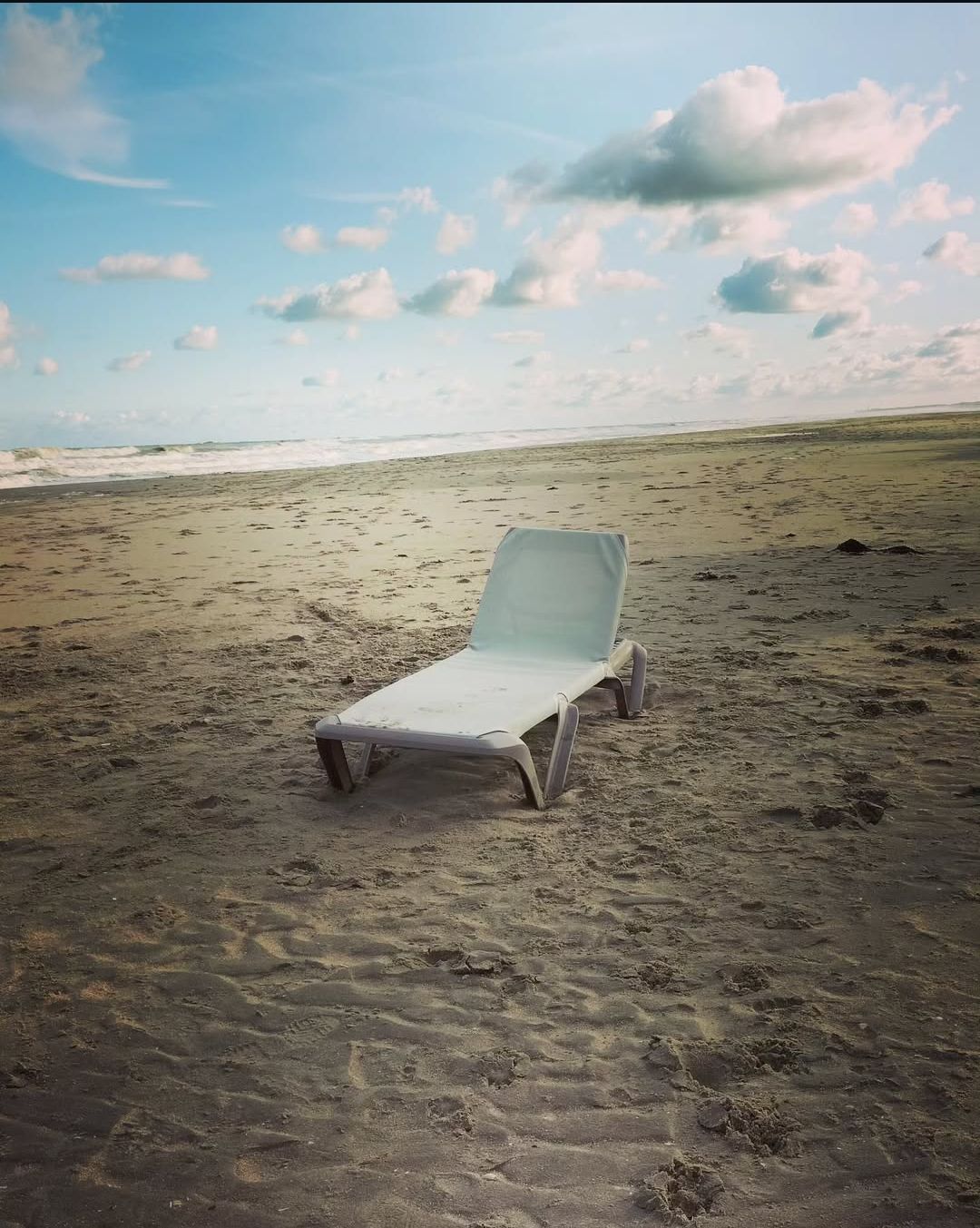 Een enkele lege ligstoel staat op een zandstrand onder een blauwe hemel met hier en daar wat wolken.