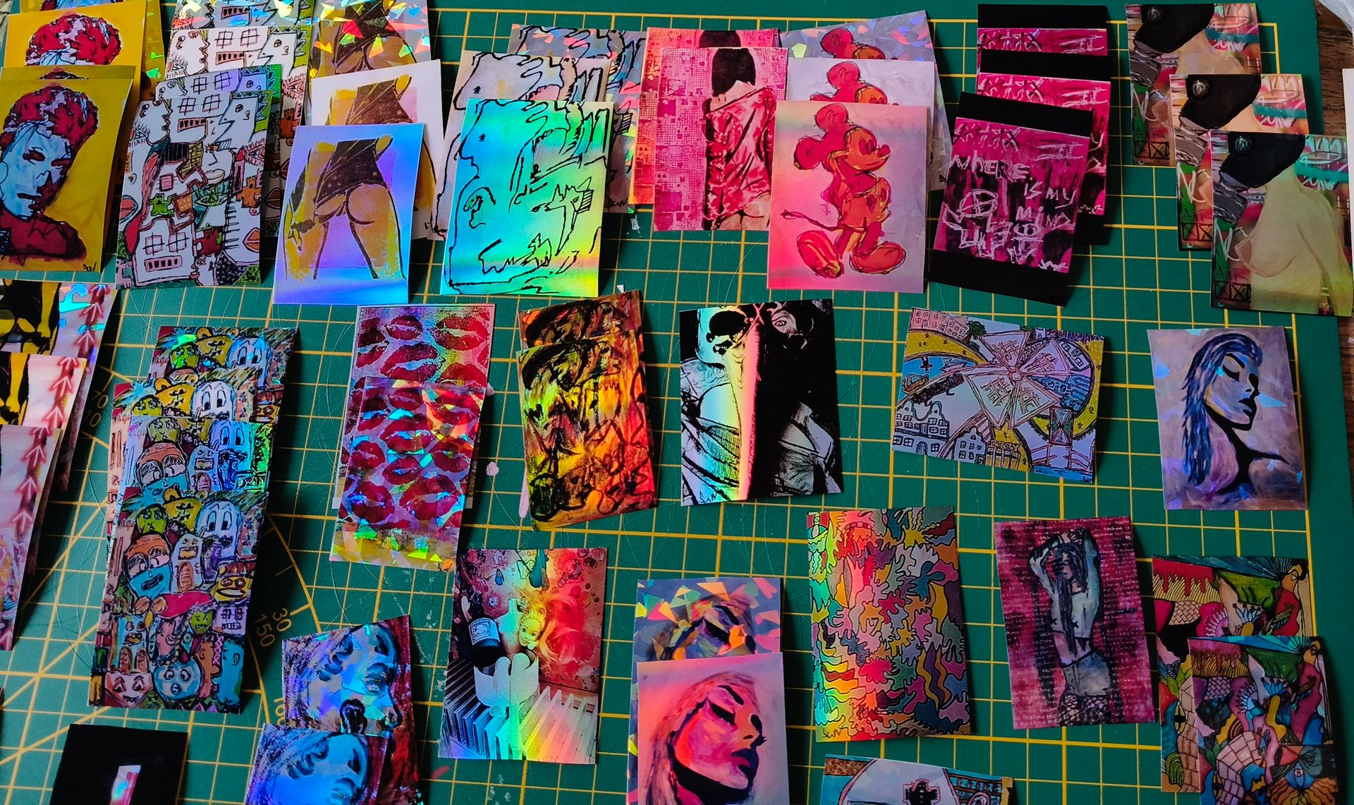 Een verzameling kleurrijke, holografische kunstprints, kaarten en stickers uitgespreid over een groene snijmat.