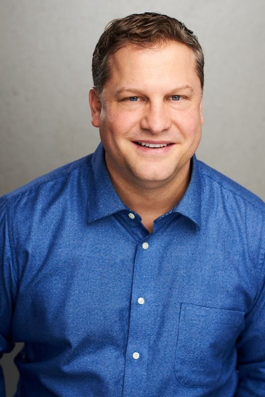 Portrait of a smiling man in a blue button-up shirt against a gray background
