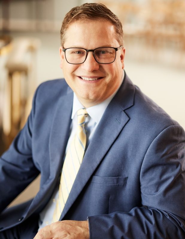 Smiling man in blue suit and glasses seated indoors against a softly blurred background