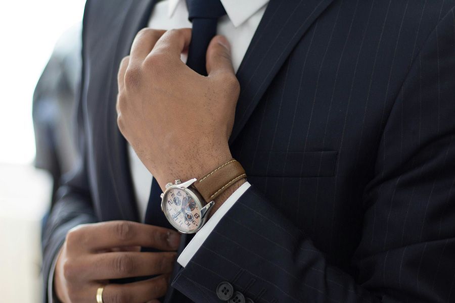 Man adjusting a dark suit and tie, wearing a wristwatch and brown leather bracelet