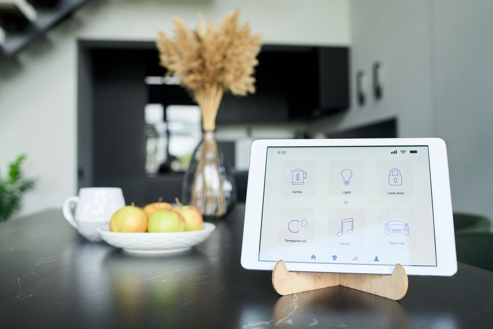 Tablet on a kitchen counter with a smart home app, beside a bowl of fruit and a vase of dried flowers.
