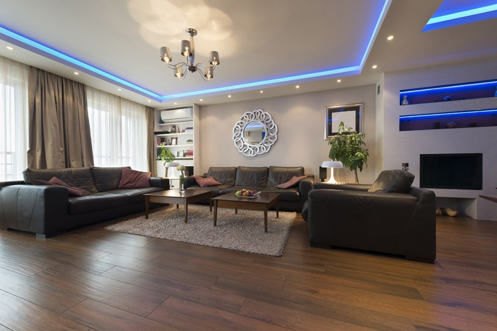 Spacious modern living room with dark sofas, wood floors, blue LED ceiling lights, and a wall-mounted TV
